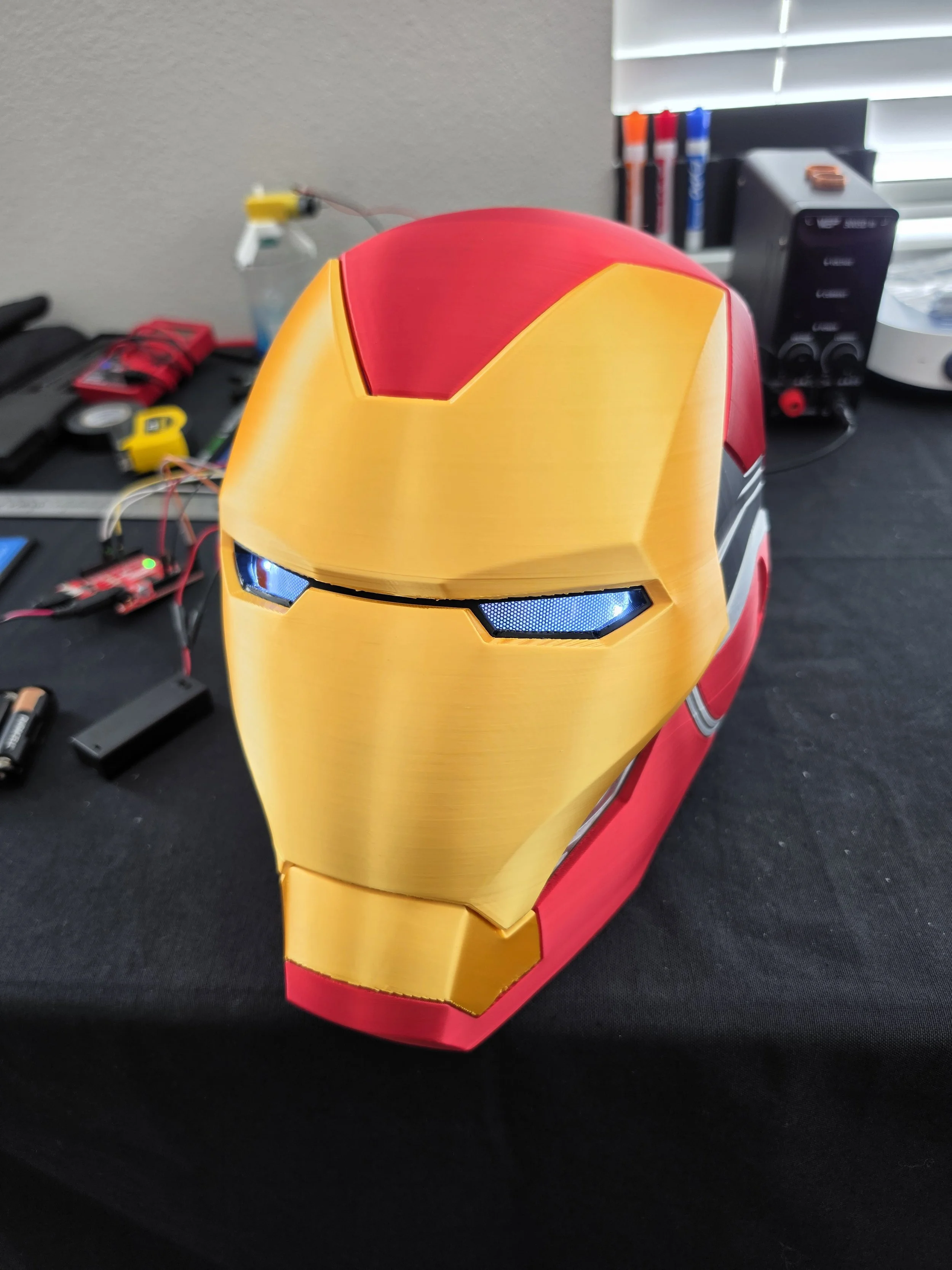 A red and yellow Iron Man helmet model on a black surface, with various electronic components and tools scattered around in the background.