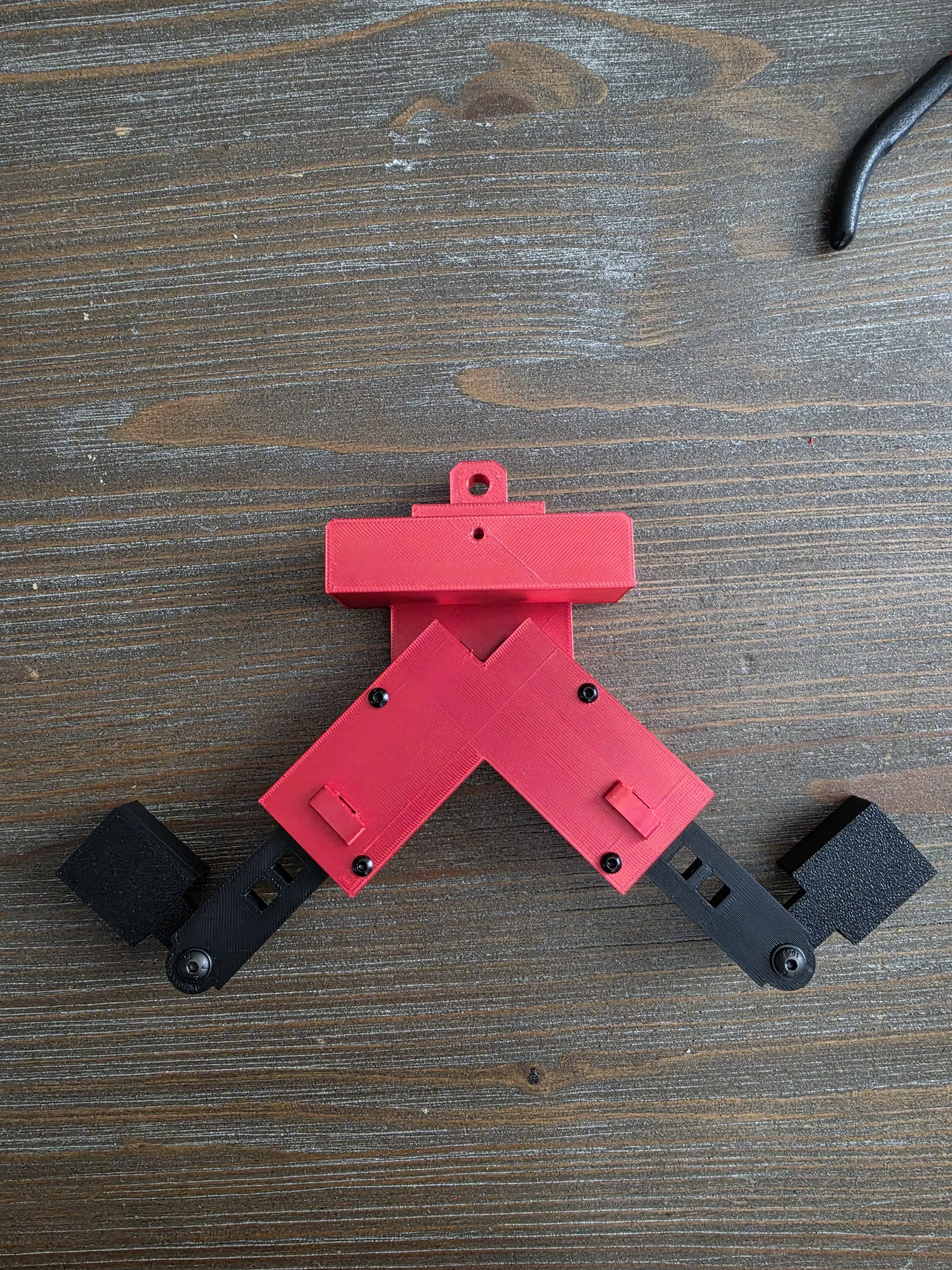 A desktop with a red 3D printed object resembling a ribbon or bow, with black straps and foam blocks attached, on a wooden surface.