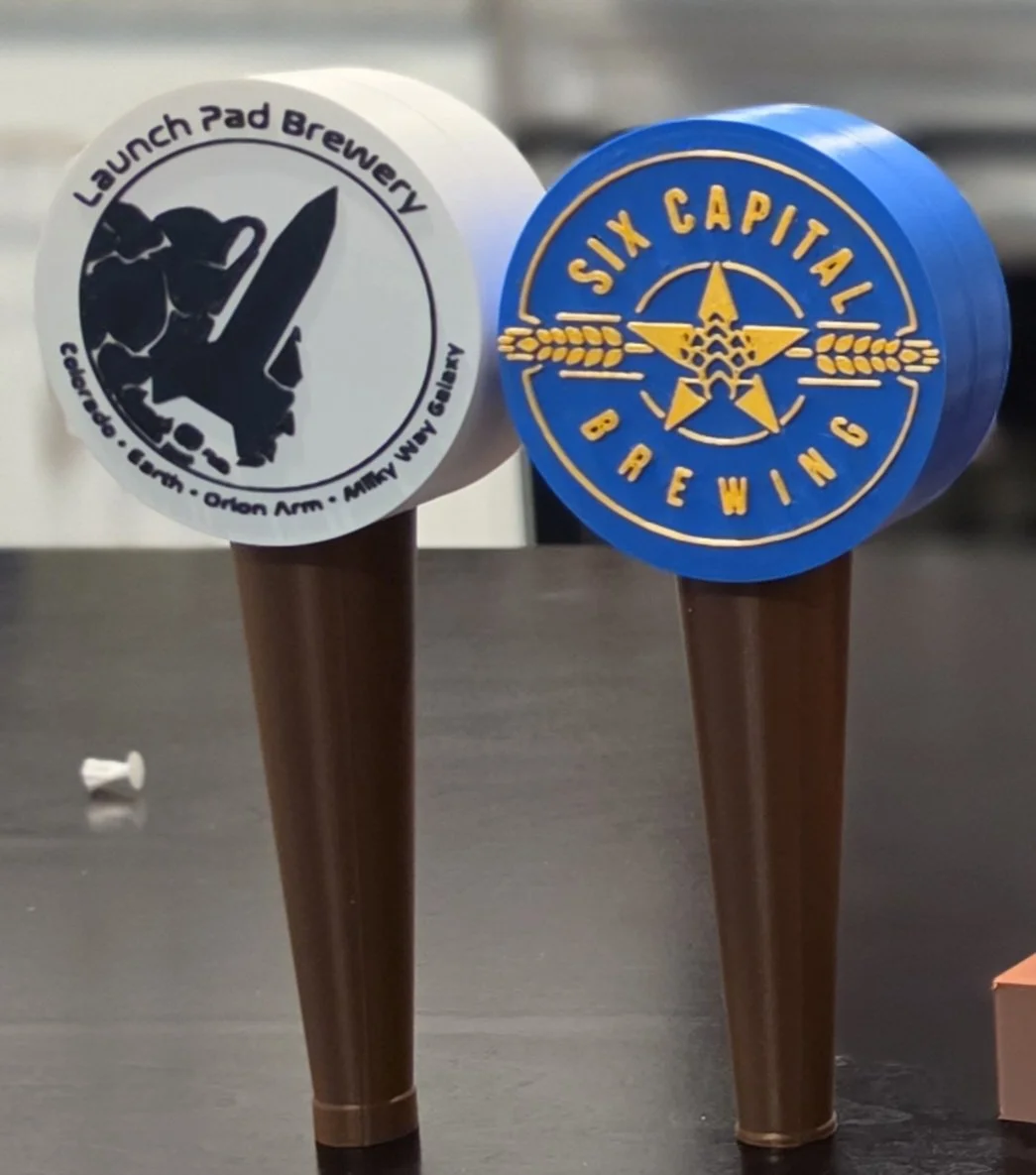 Two beer taps with branding logos; one for Launch Pad Brewery with a space shuttle graphic, and the other for Six Capital Brewing with a star and wheat graphic.