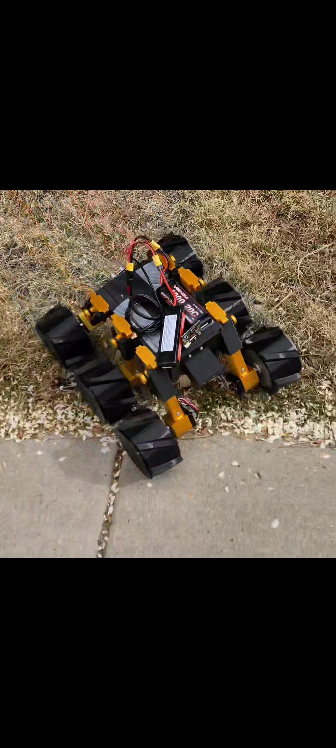 A small tracked robot with black and yellow parts on a sidewalk next to a patch of grass and dried leaves.