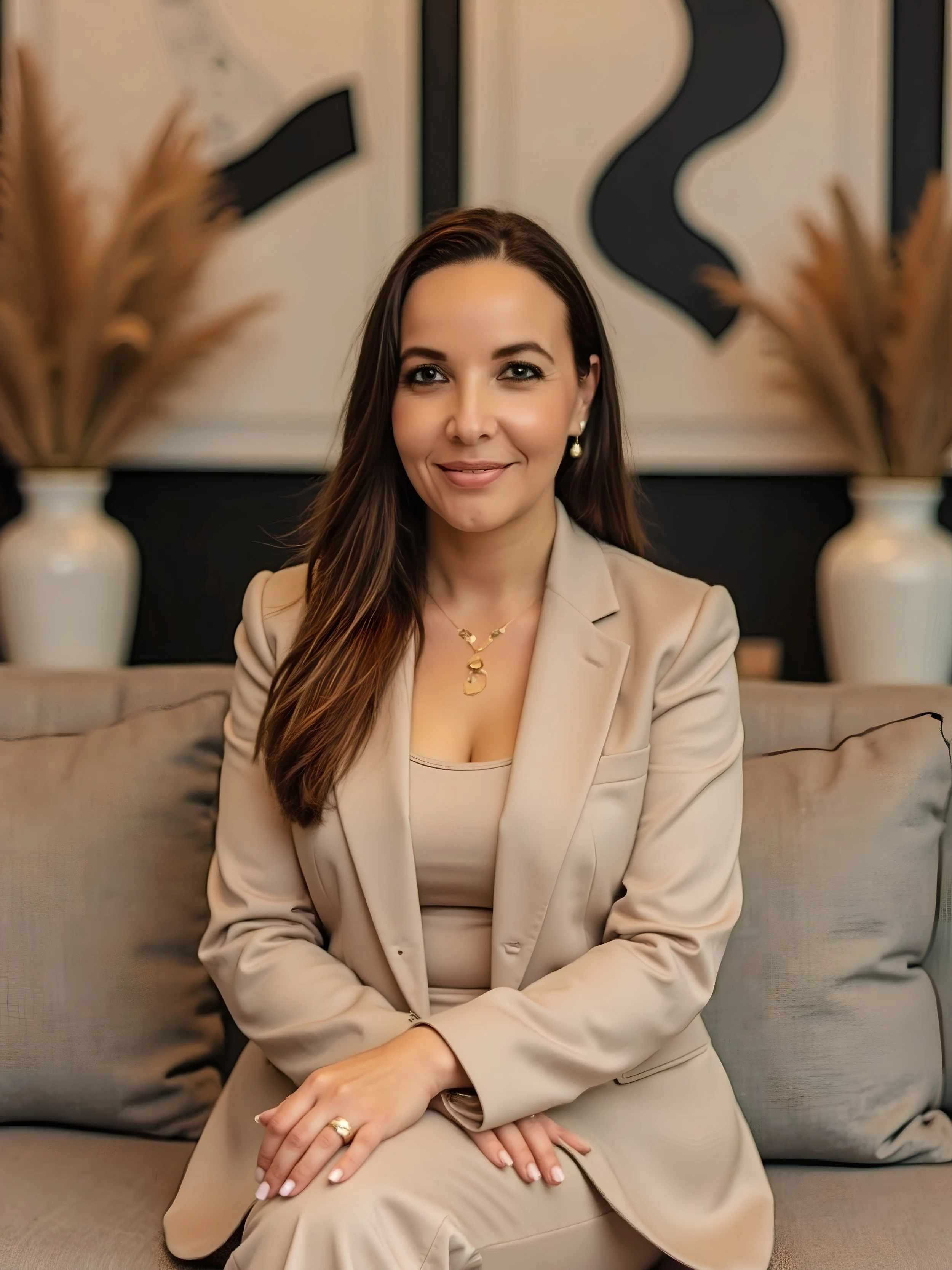 Una mujer joven con cabello largo, vestida con un traje beige, sentada en un sillón beige, con fondo de paredes blancas y decoraciones en negro y beige.
