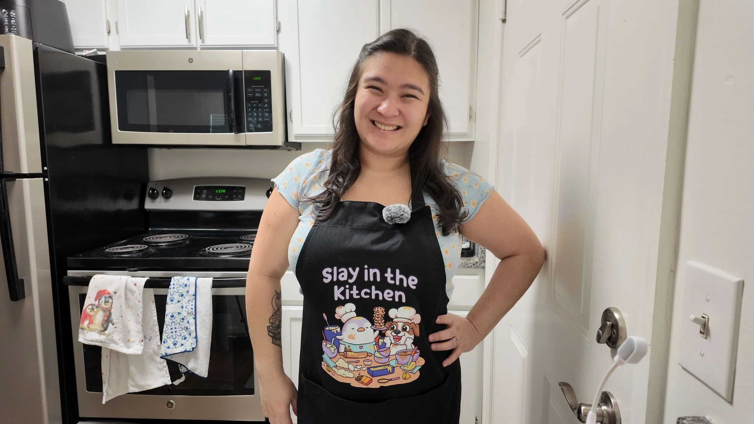 A woman standing in a kitchen, smiling, wearing a black apron that says 'slay in the kitchen' with cartoon characters and baked goods. The kitchen has white cabinets, a microwave, an oven, and dish towels hanging on the oven handle.