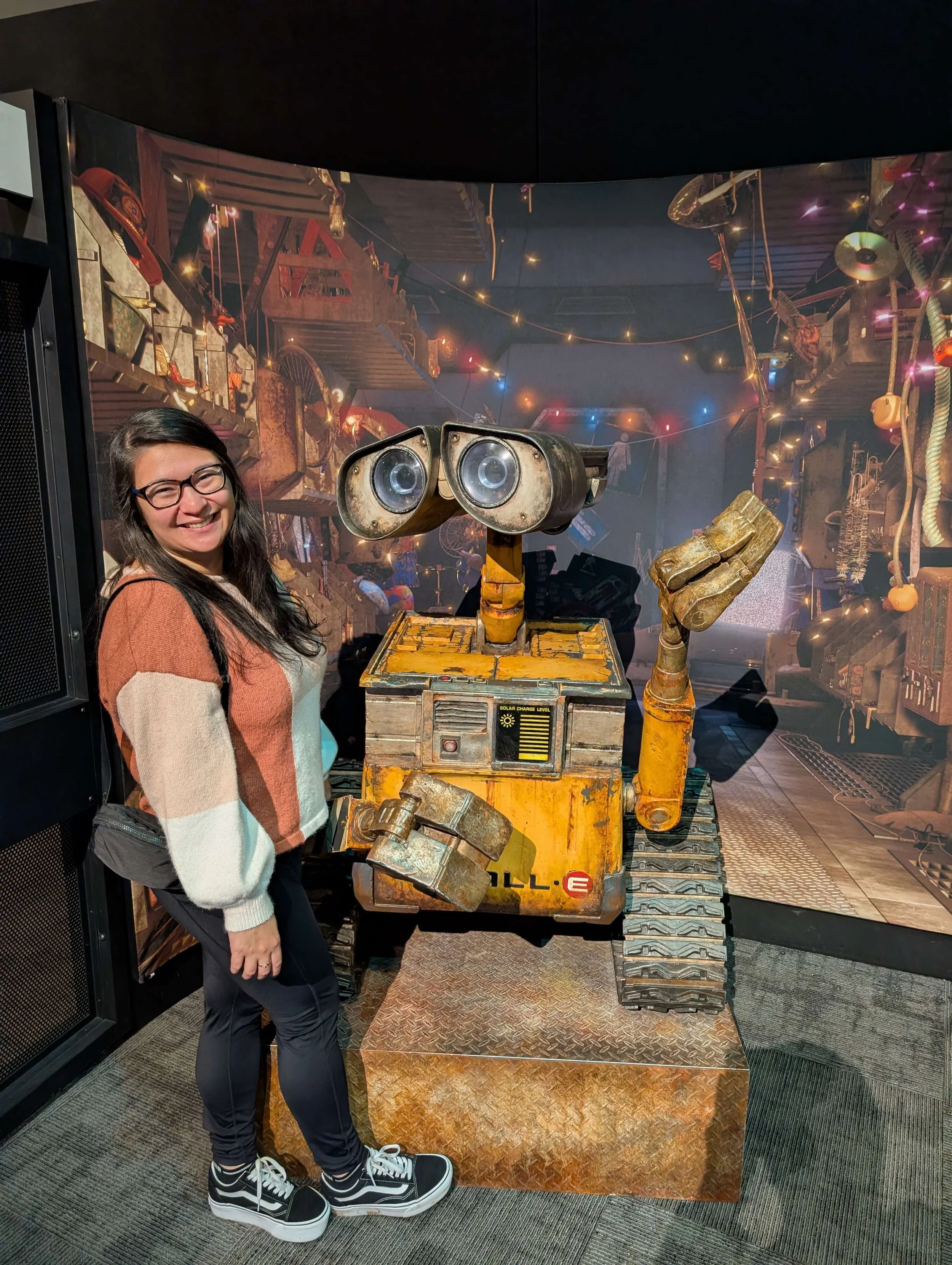 A woman with glasses, long dark hair, wearing a multicolored sweater and black pants, smiling and standing next to WALL-E, a robot with tank-like treads, a boxy yellow body, and binocular-like eyes, in front of a decorated backdrop with warm string lights.