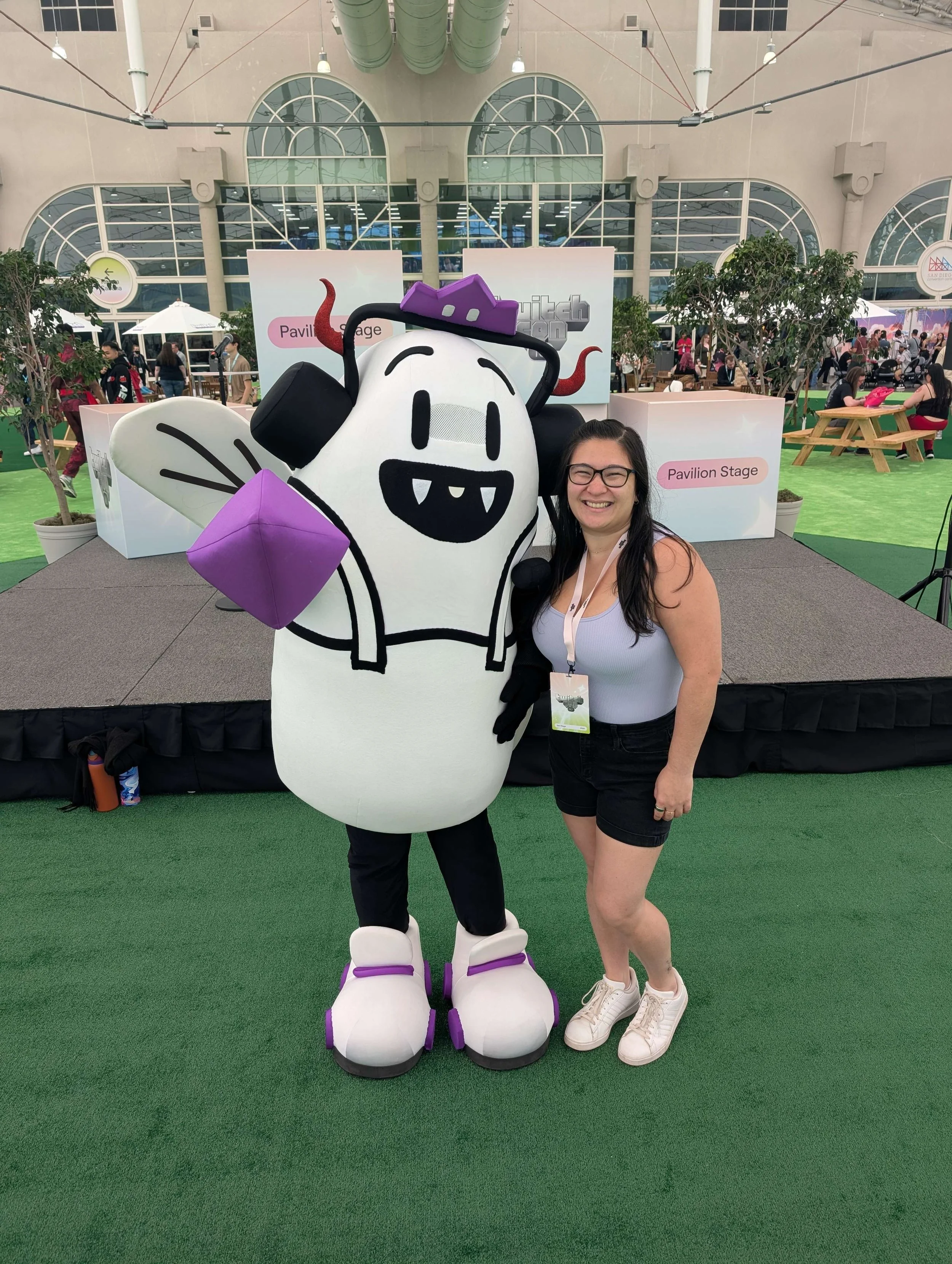 A woman with long dark hair, glasses, and wearing a white tank top and black shorts, smiling at the camera, stands beside a mascot dressed as a cartoon character with a white head, black markings, purple hat, black and purple accessories, and large white shoes with purple accents, on a stage at an indoor event with people in the background and signages indicating a Pavilion Stage.
