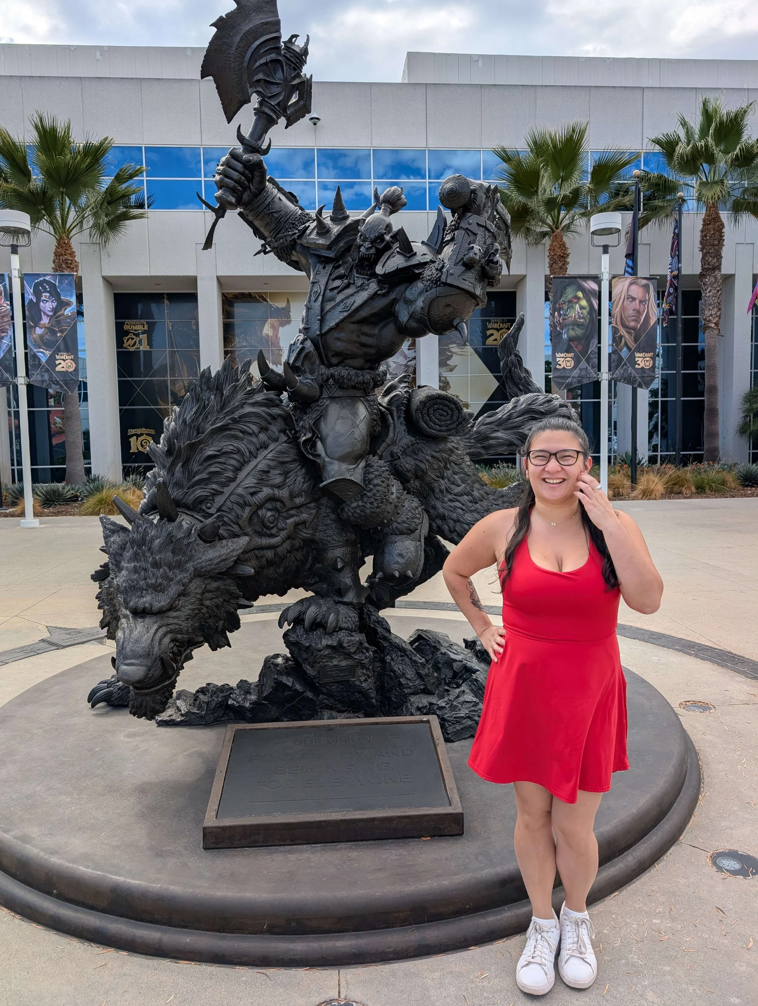 A woman in a red dress and white sneakers standing in front of a black statue of a warrior riding a creature, outside a building with palm trees and banners for the game World of Warcraft in the background.