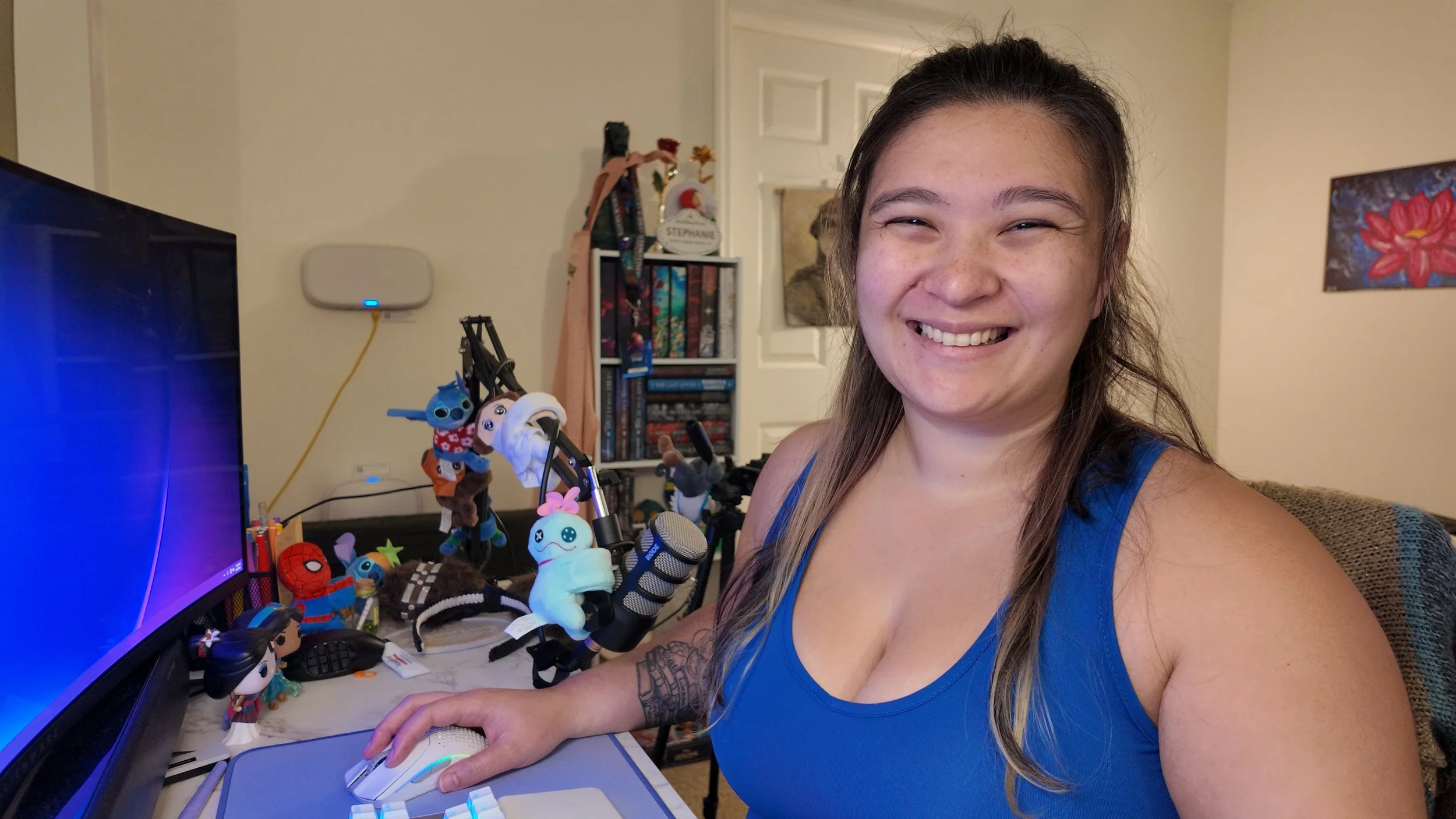 A woman with long hair smiling at a camera, sitting at a desk with a computer and surrounded by plush toys, action figures, and gaming accessories.