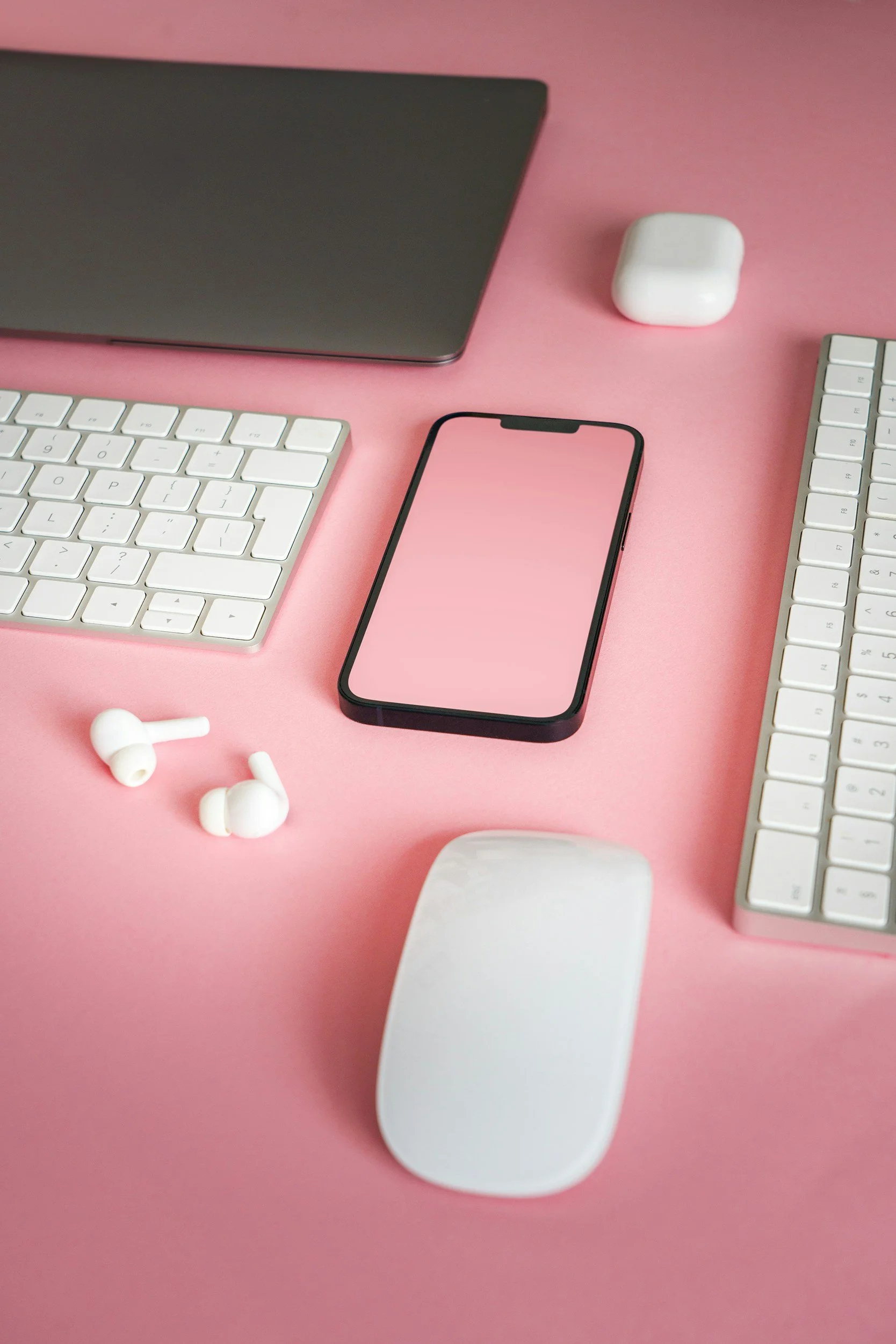 A pink desk with wireless earbuds, a smartphone, a wireless keyboard, a computer monitor, a wireless mouse, a white AirPod case, and two white keyboard cases.