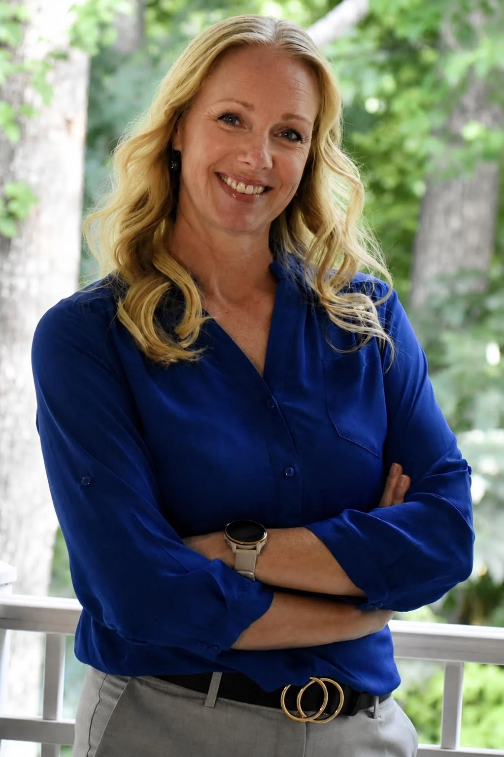 A woman with blonde hair, wearing a blue blouse and gray pants, smiling and standing outdoors with arms crossed.