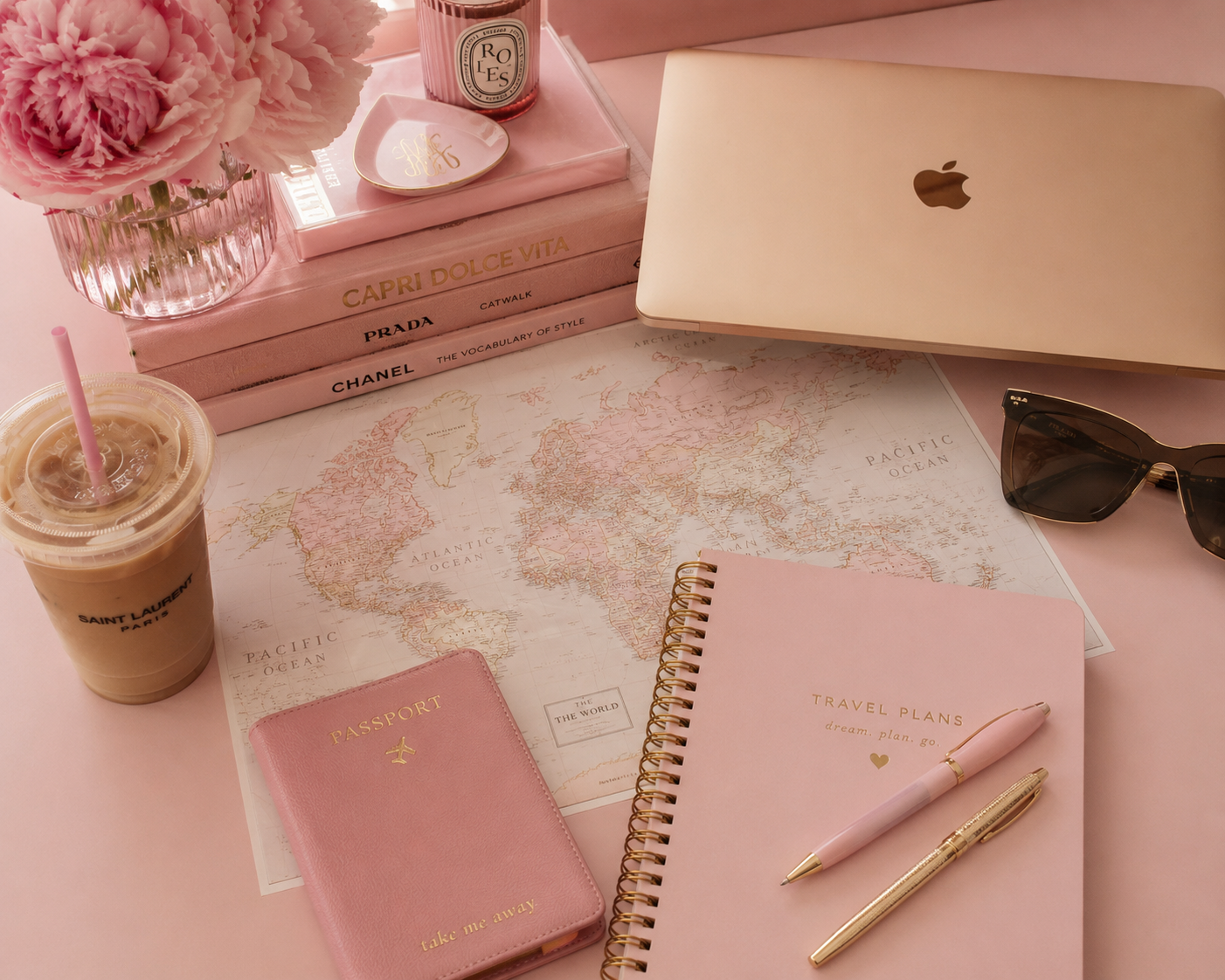 A flat lay of travel essentials and decor featuring a world map, pink books, a laptop, sunglasses, a pink passport holder, a pink travel planner, two pens, a coffee cup, a candle, flowers, and a small dish on a pink surface.