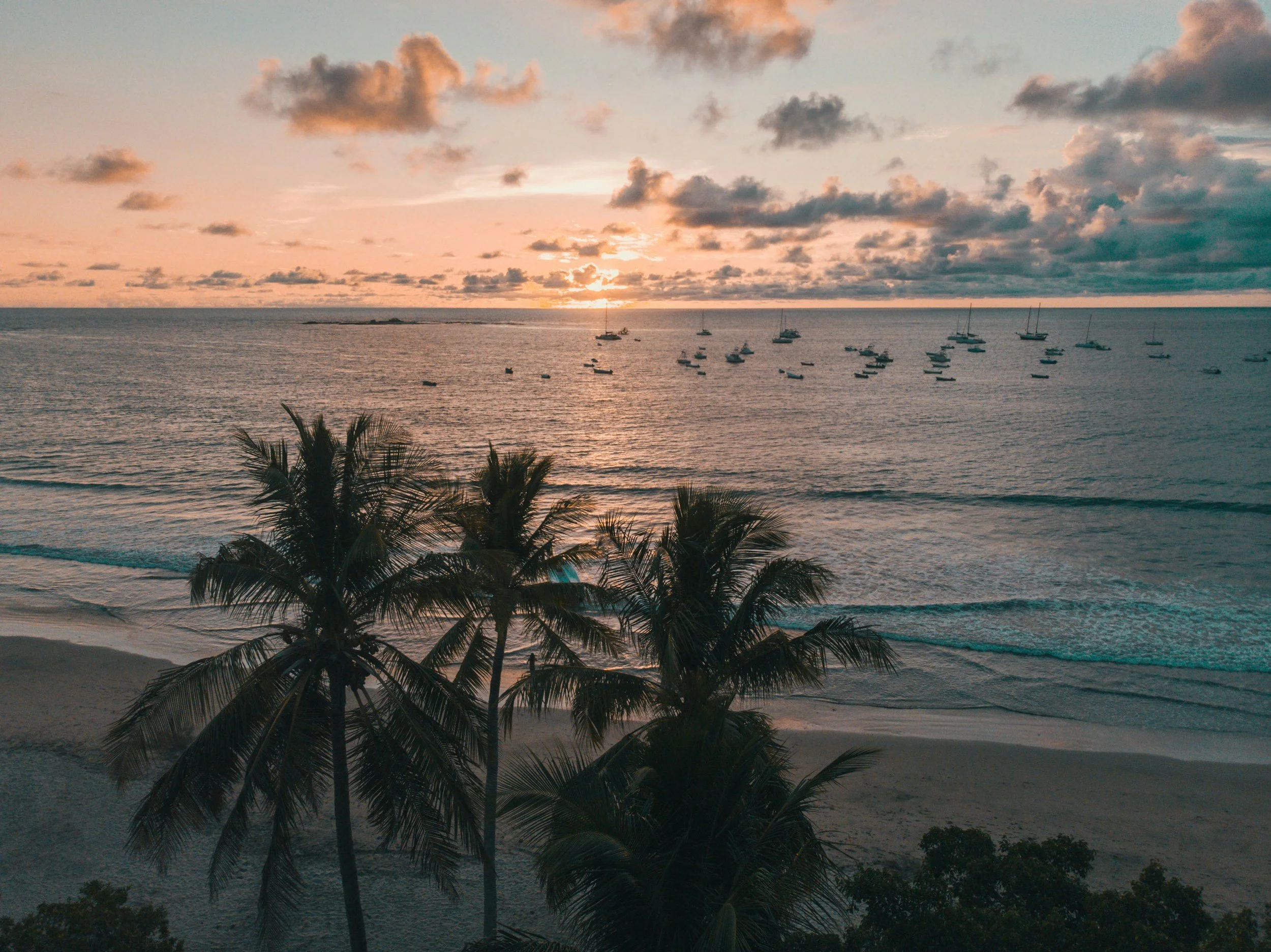 A sunset over the ocean with several boats anchored, palm trees in the foreground, and a partly cloudy sky. Costa Rica Beach.