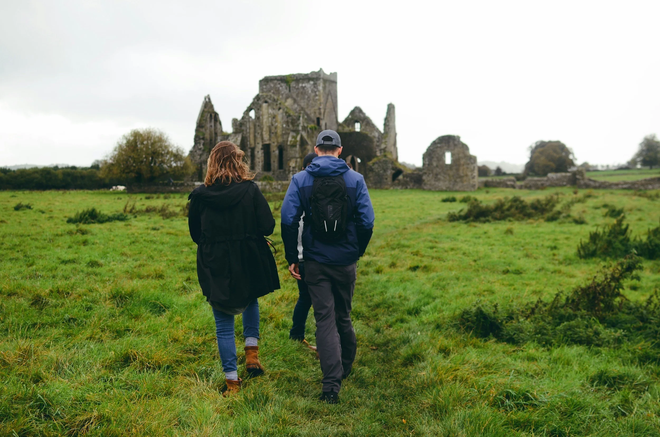 Three people walking through a grassy field with ruins of an old stone castle in Ireland in the background on a cloudy day.