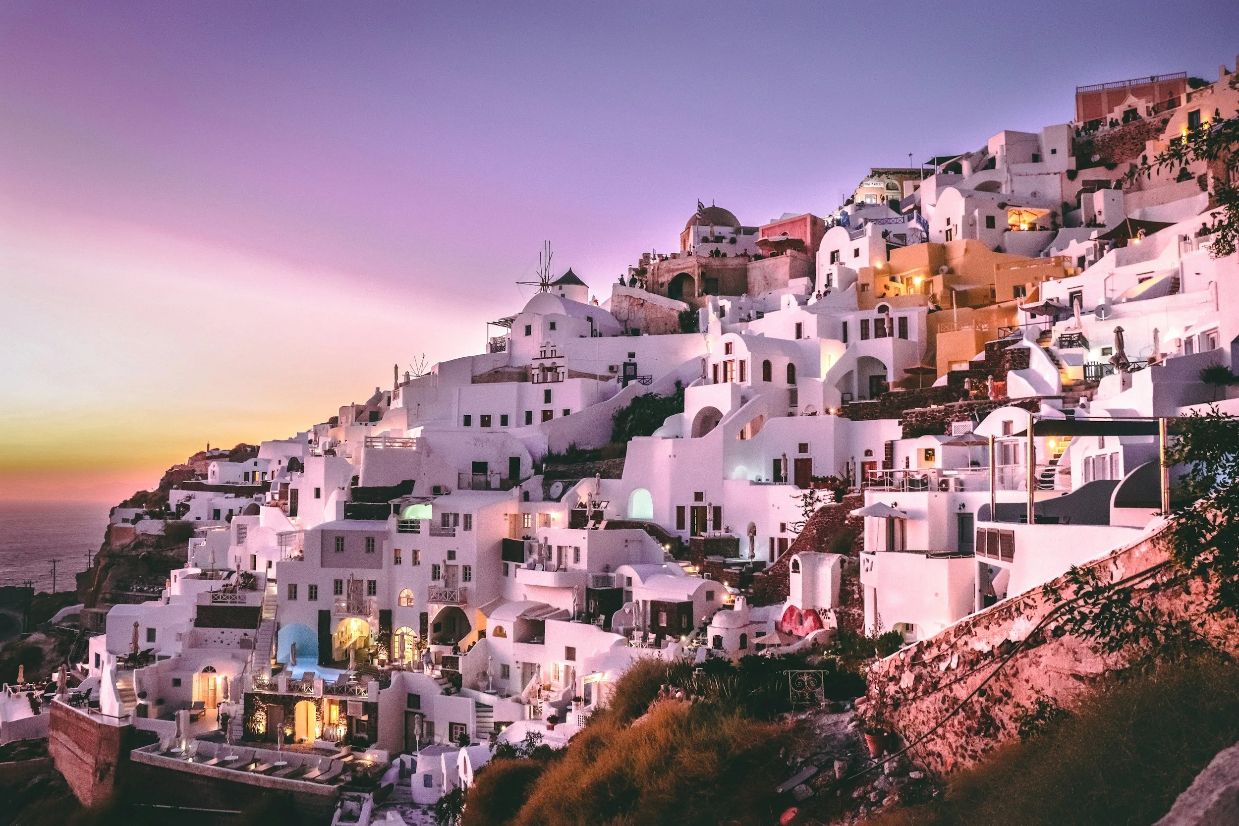 White-washed buildings with domed roofs and terraces built on a hillside at sunset, typical of Santorini, Greece.