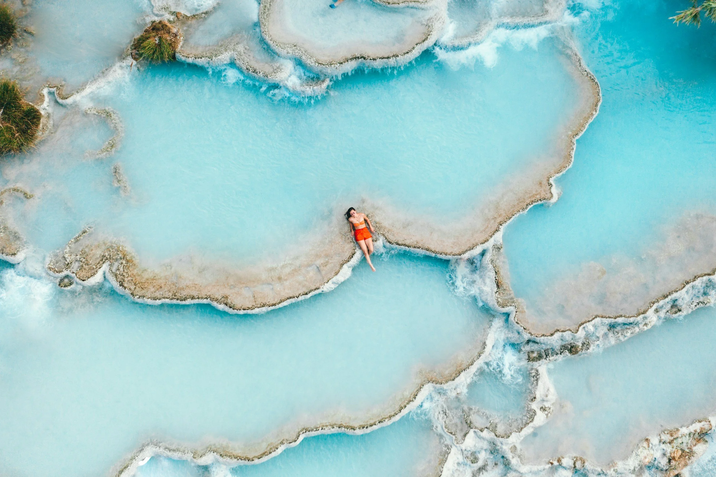 A woman lying on the edge of a natural hot spring pool with layered mineral deposits, surrounded by water and greenery.