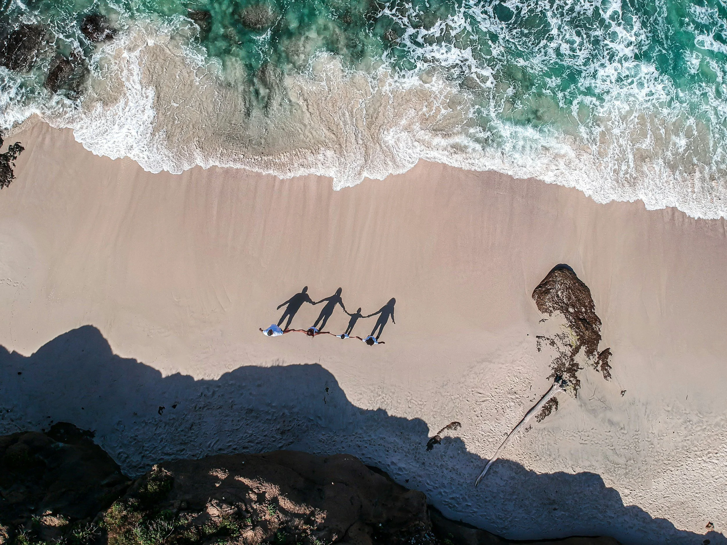 Bird's eye view of beach with turquoise water and foamy waves crashing onto sandy shore. Four people hold hands on the beach, their bodies casting shadows on the sand as the ocean waves meet their feet.