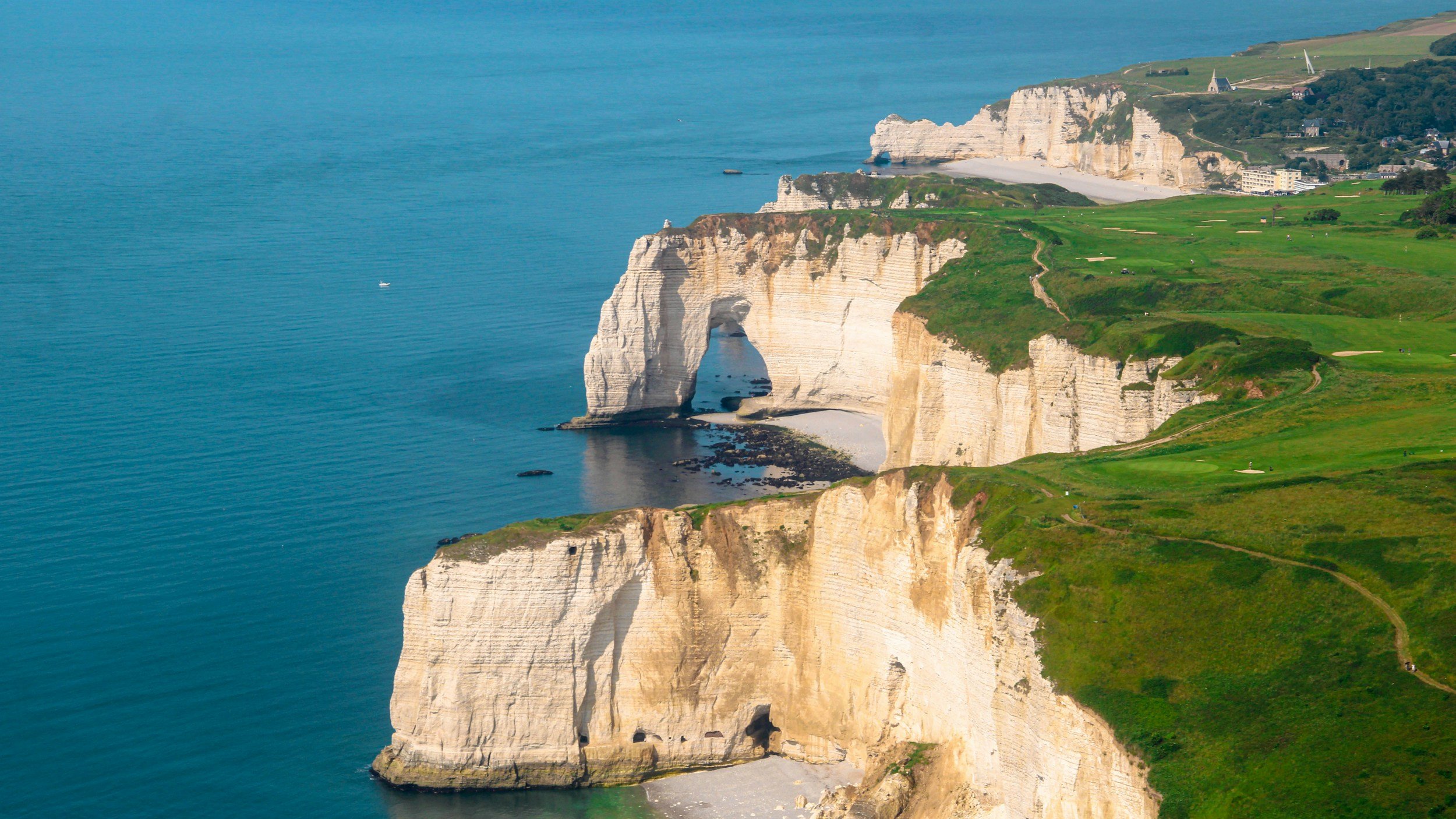 Cliff side view of white chalk cliffs with a natural arch, green grass terrain, small beach, and a village in the background by the ocean. Golf course in Scotland.