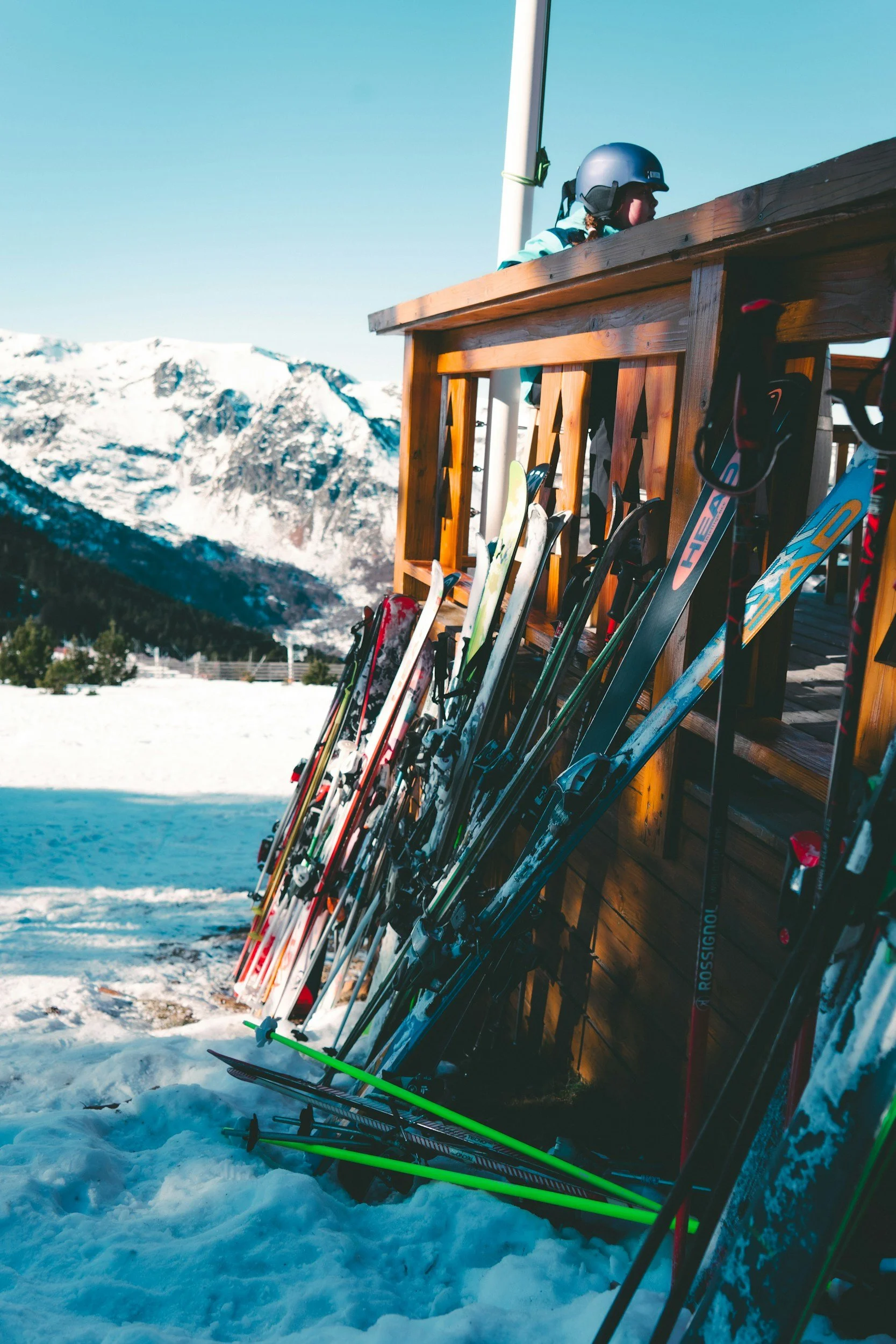 Skis and ski poles leaning against a wooden rack outside in the snow, with snowy mountain peaks and a clear blue sky in the background.