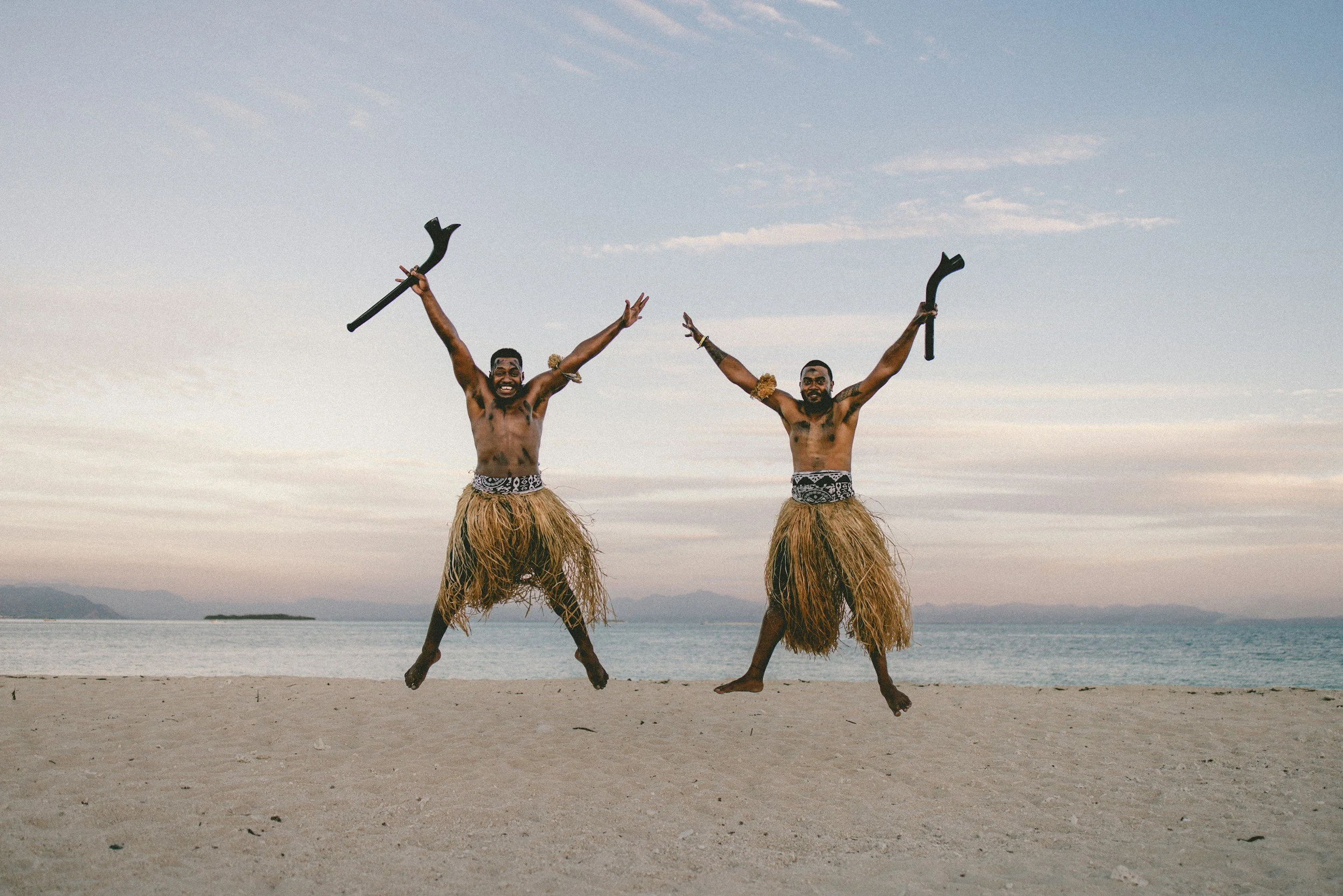Two men with traditional grass skirts jumping in the air on a sandy beach, holding black weapons, with a vast ocean and mountains in the background. Fiji.