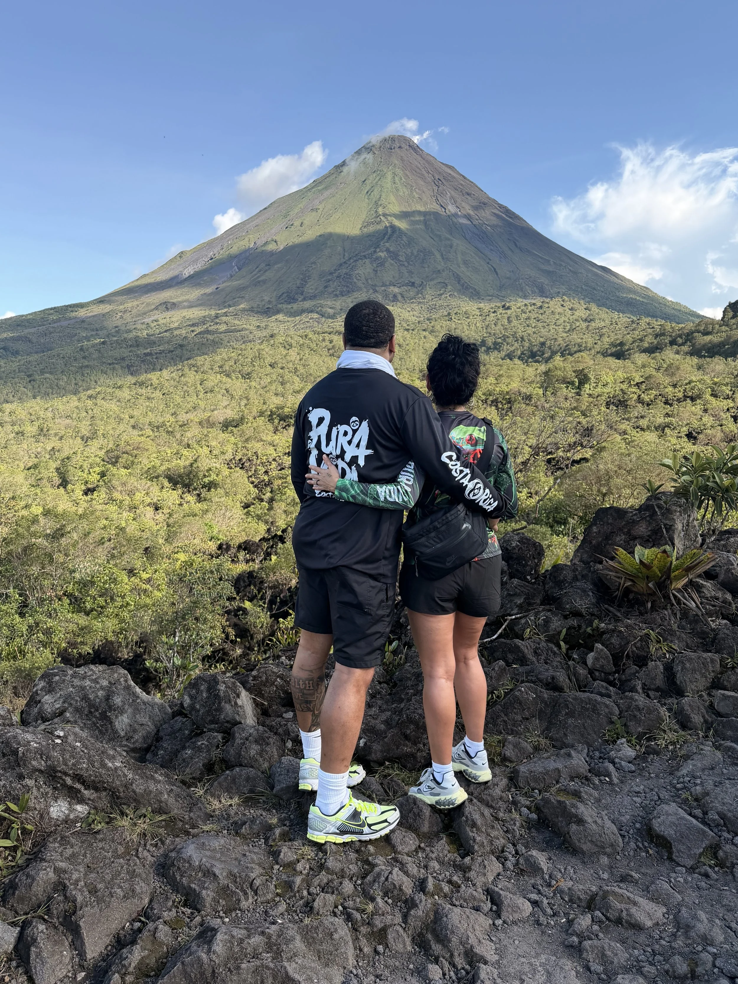 Two people standing on rocky terrain, gazing at Arenal Volcano with a conical shape and smoke coming out from the summit, in a lush green landscape under a blue sky with some clouds.