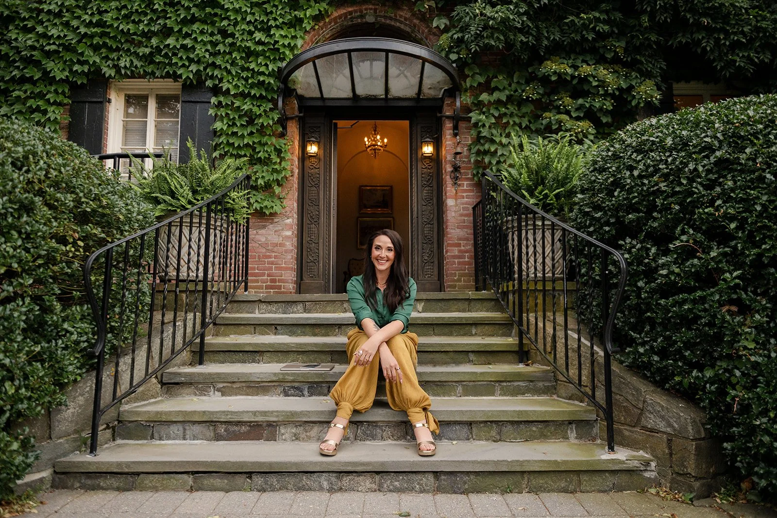 A woman sitting on the steps of a brick estate entrance surrounded by green bushes and ivy, smiling at the camera. Looks to be somewhere in Europe or New England.