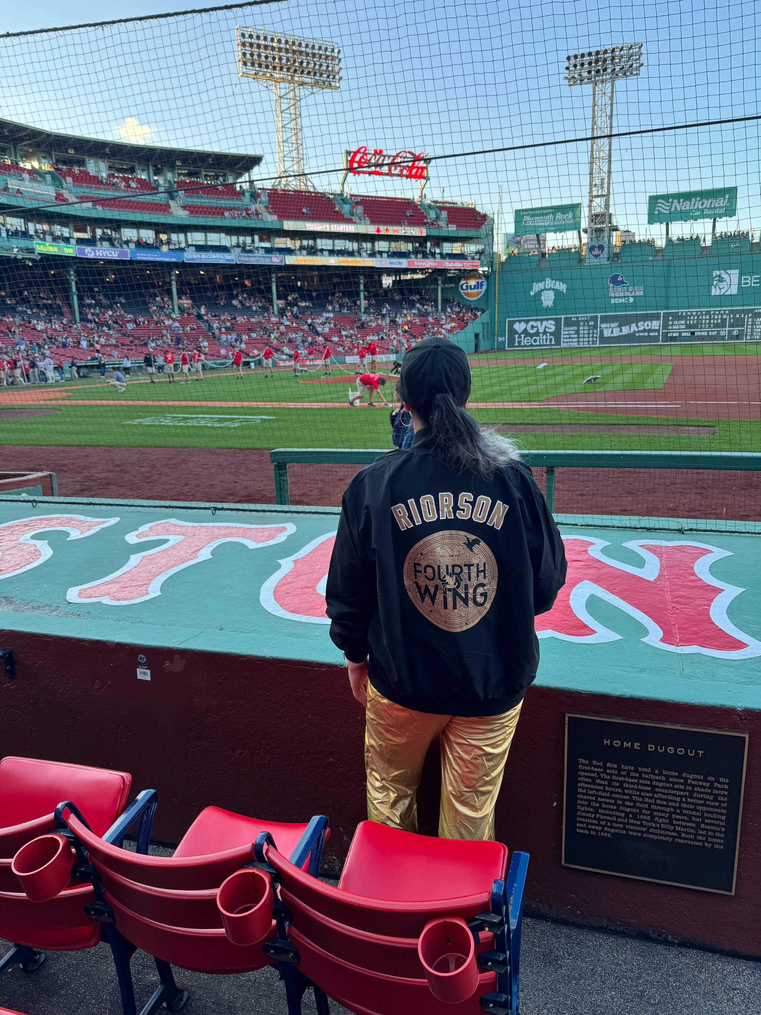 A  wearing a blackjacket with 'Riorrson Fourth Wing' written on the back observes a baseball game from behind a safety net. Fenway Park, Boston, MA. Players are warming up on the field, and a large scoreboard is visible in the background.