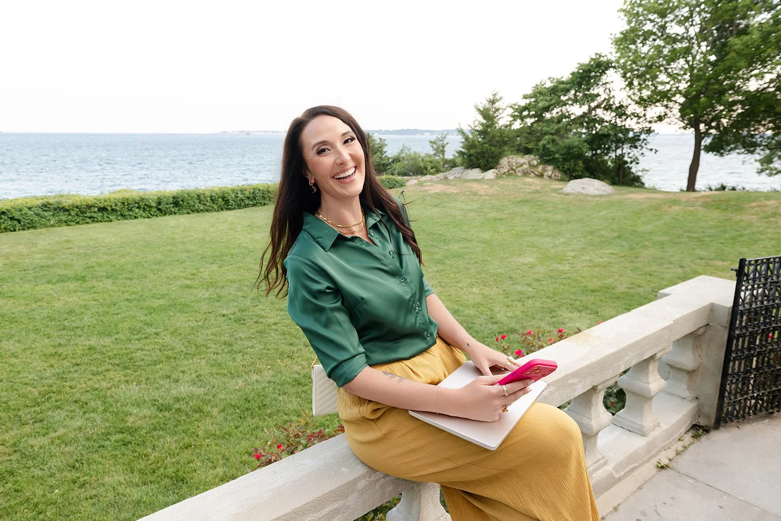 A woman sitting on a stone bench outdoors near a body of water, holding a pink phone and a white notebook, smiling at the camera. She is wearing a green blouse and yellow pants, with trees, rocks, and the ocean in the background.