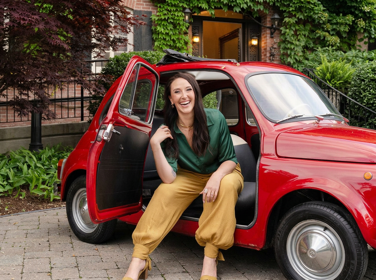A woman with long dark hair, wearing a green blouse and mustard yellow pants, sits on the open door of a vintage red Fiat, smiling surrounded by greenery and a brick estate entrance.