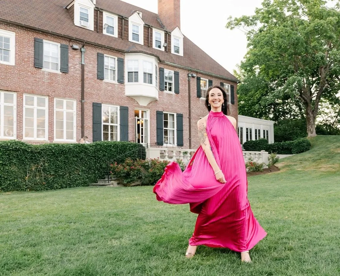 A woman in a bright pink dress dancing on a lawn in front of a brick estate with black shutters and multiple windows. Located in Beverly, MA, USA.