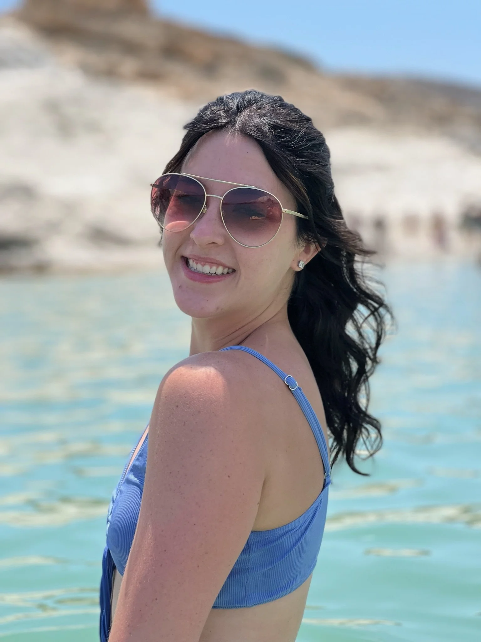 A woman with dark, wavy hair wearing sunglasses and a blue swimsuit, smiling at the camera, on a sandy beach with clear water and a blurred background of sand dunes and sky. Moon Beach, Milos, Greece.