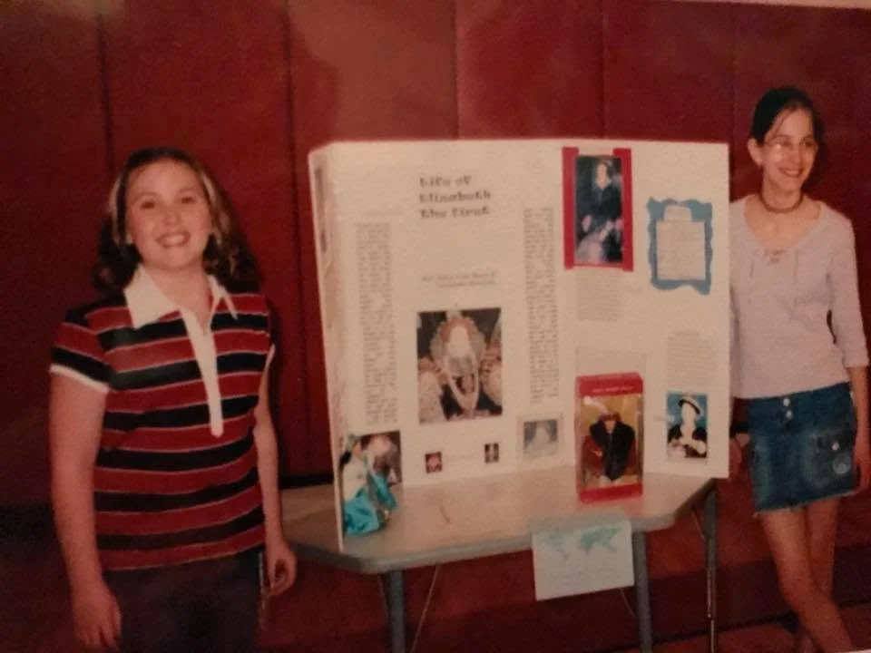 Two young girls standing next to a science project display board, smiling indoors with wood-paneled walls. British Monarch presenation.