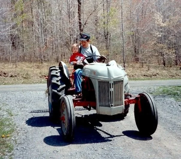 A man and a young girl sitting on an old tractor on a gravel road with a wooded background during daytime.