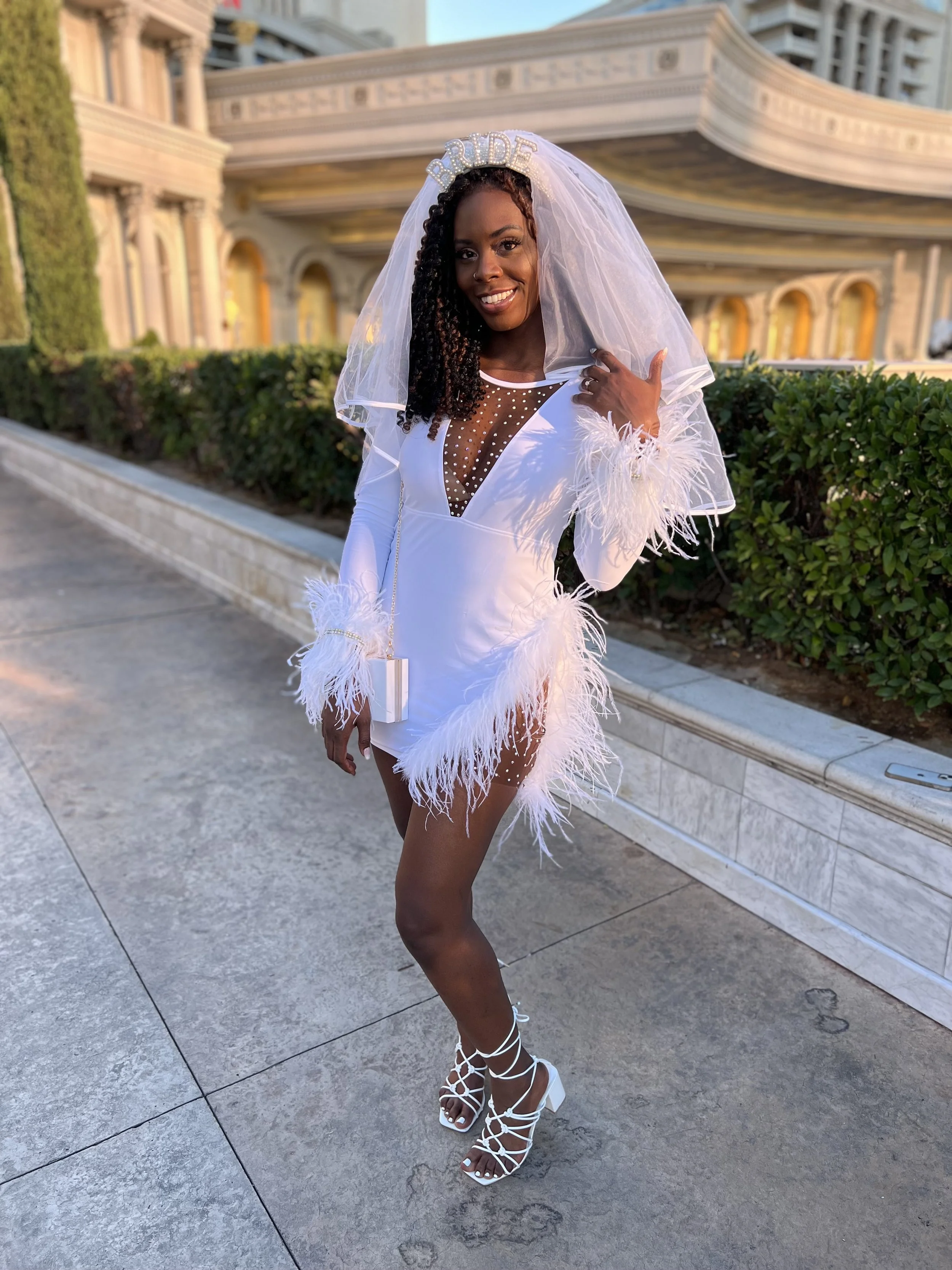 A woman dressed in a white dress with feathered trim and a veil that says 'BRIDE' in rhinestones, standing outside on a stone walkway with green bushes and a large ornate building in the background, smiling and posing for the photo. Las Vegas, NV.
