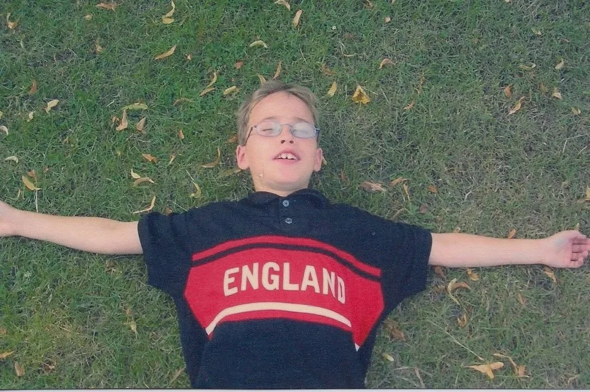 Twelve year old boy laying in the grass with his arms sprawled out on his sides wearing a black shirt with a red stripe across the front with white block lettering that says England.