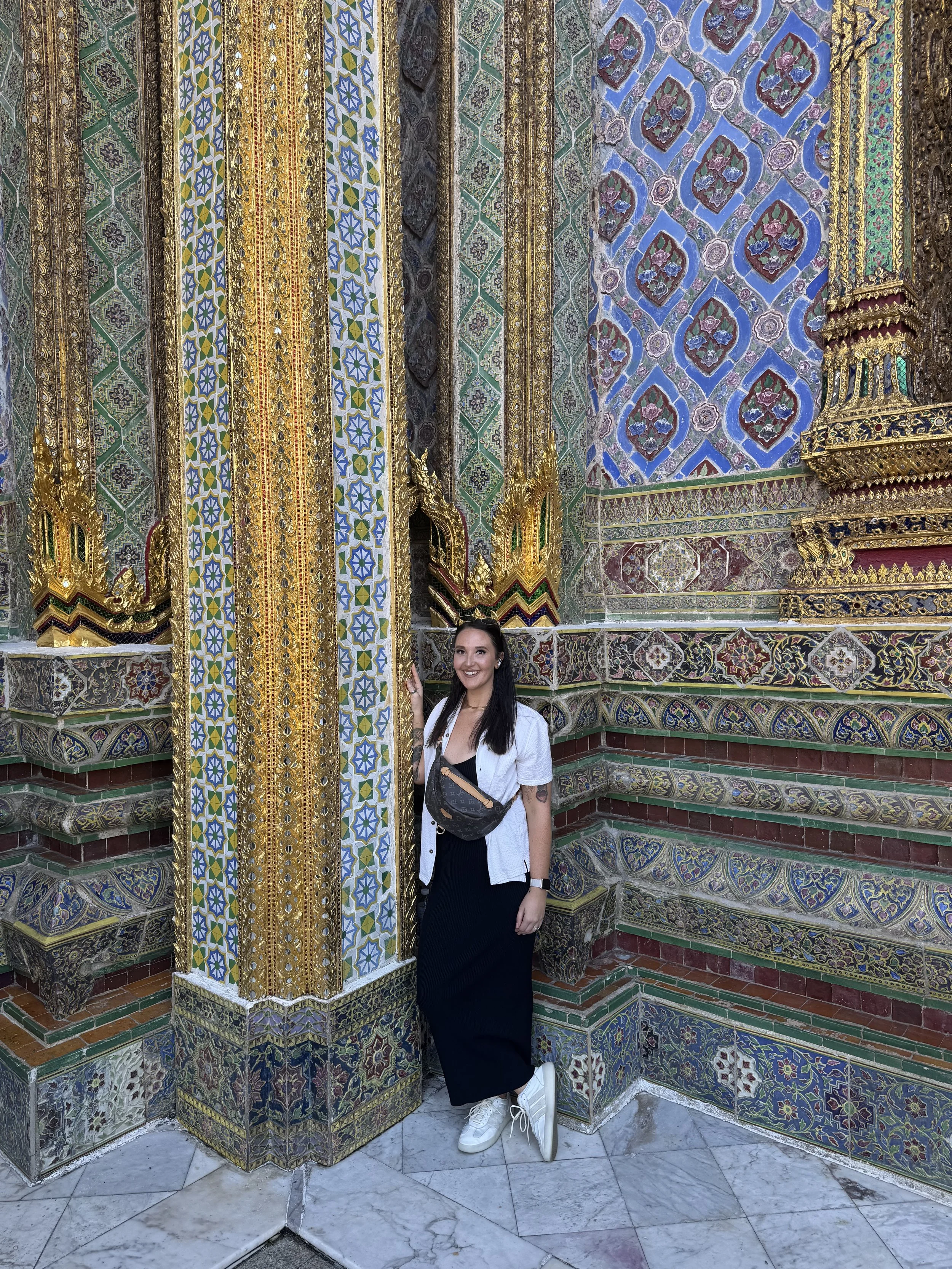 A woman with long dark hair wearing a black dress, white sneakers, a white cardigan, and a Louis Vuitton Bum Bag is standing against a wall decorated with colorful, ornate mosaic tiles and gold accents, at a temple in Thailand.