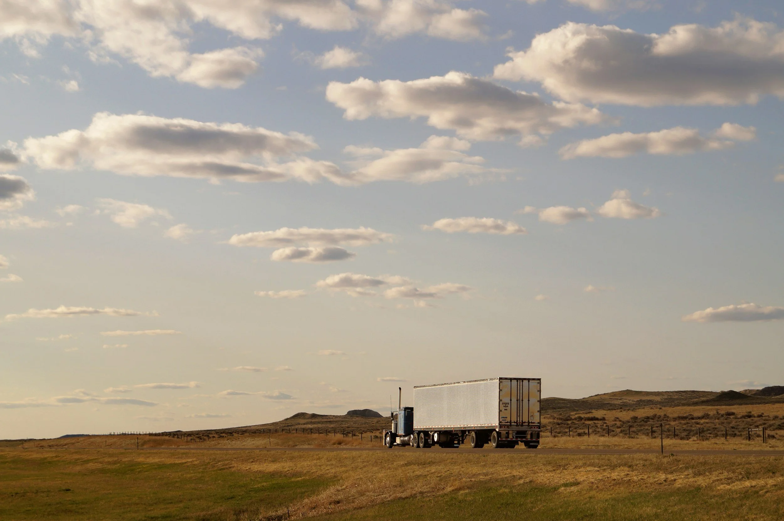 A large semi-truck driving on open road in a rural landscape with grassy fields, hills, and a partly cloudy sky.