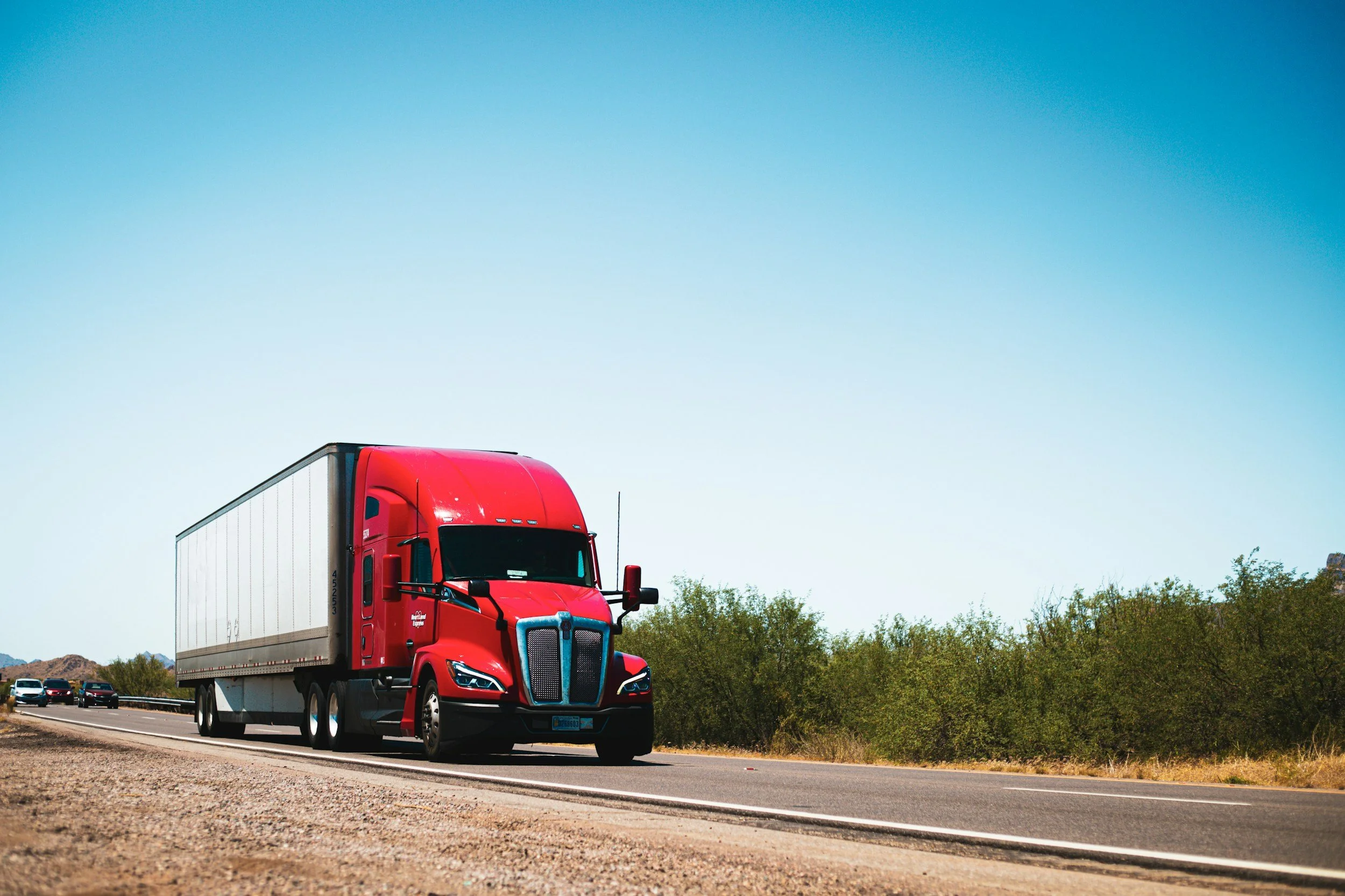 Red semi-truck driving on a highway with green trees and a blue sky in the background.