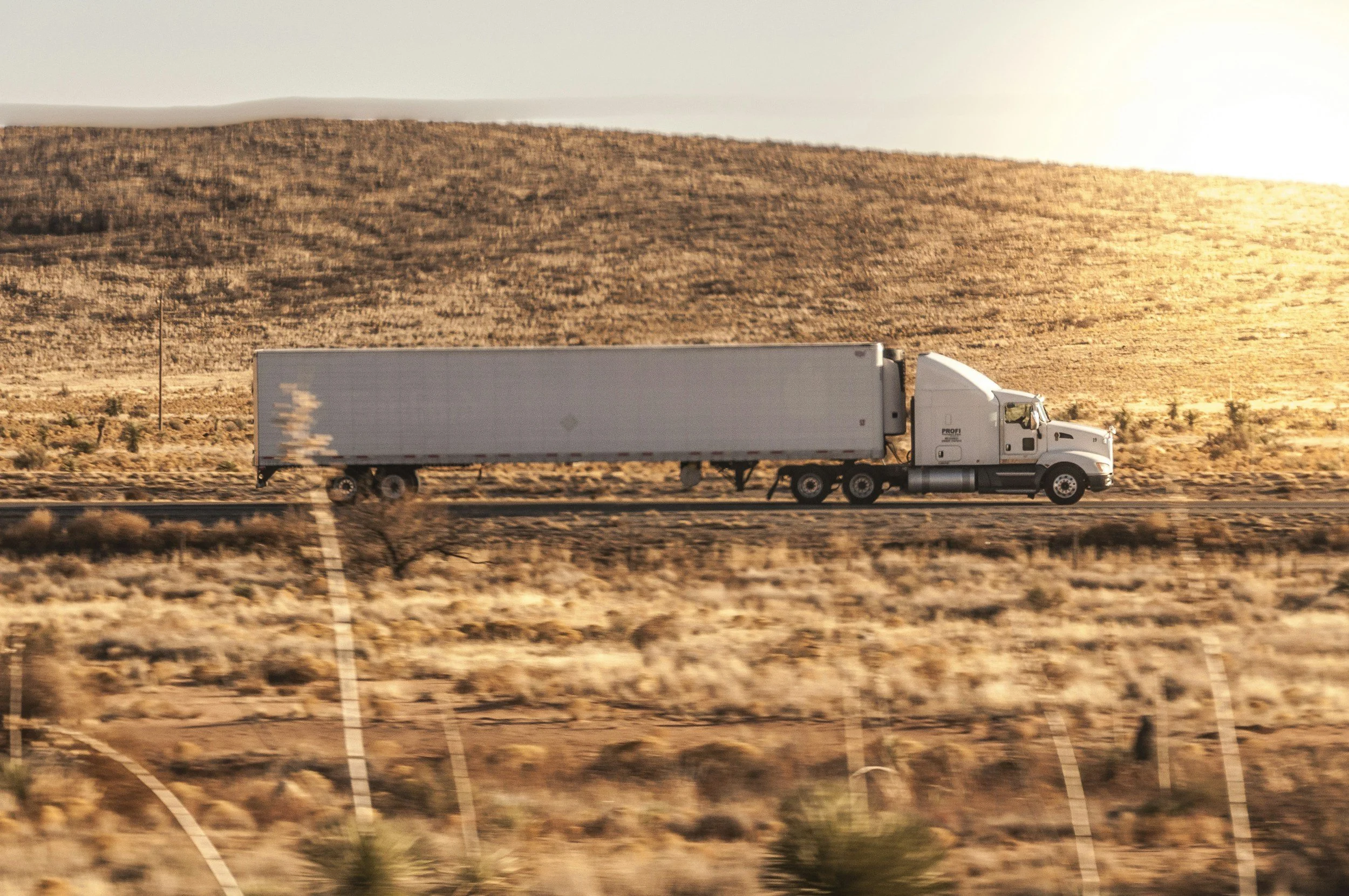 A white semi-truck driving on a highway through a desert landscape with hills in the background during sunset.