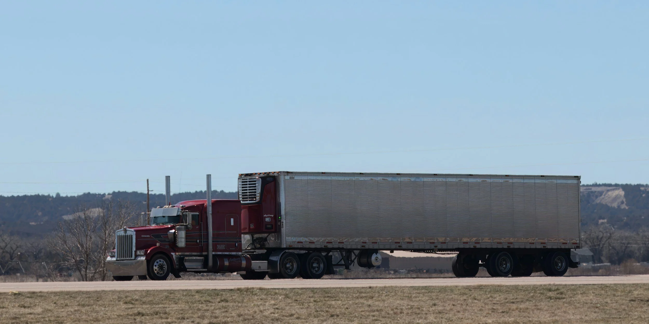 A red semi-truck with a silver trailer driving on a highway with trees and hills in the background under a clear blue sky.