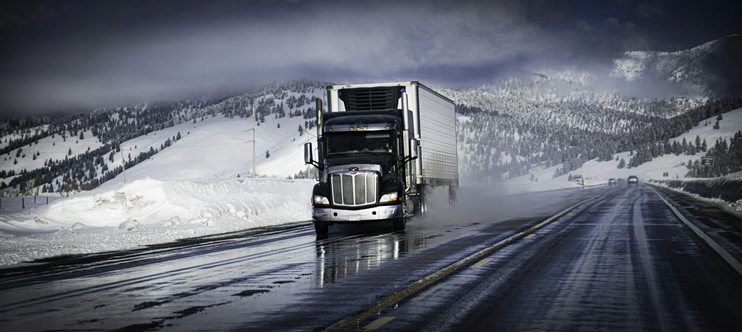 A black semi truck driving on a snowy mountain highway with snow-covered trees and mountains in the background.