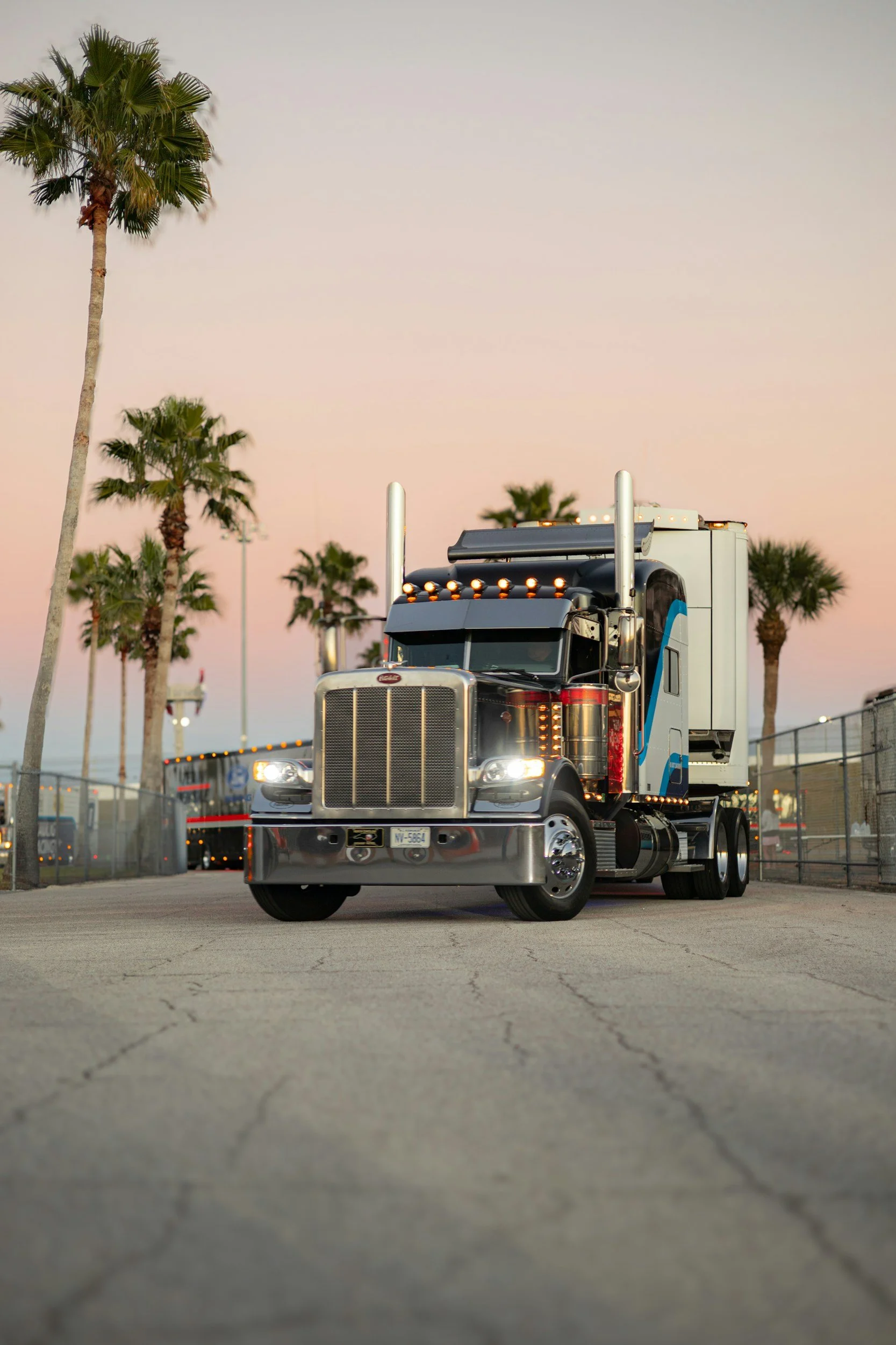 A semi-truck with a black grille and silver accents driving on a paved road, with palm trees and a chain-link fence in the background during dusk or dawn.