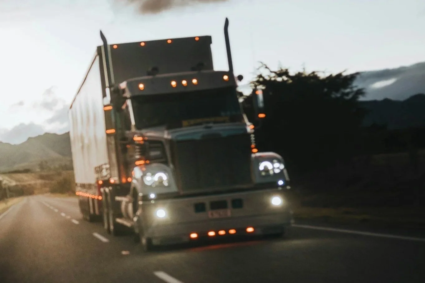 A large black semi-truck driving on a highway at dusk with mountains and cloudy sky in the background.