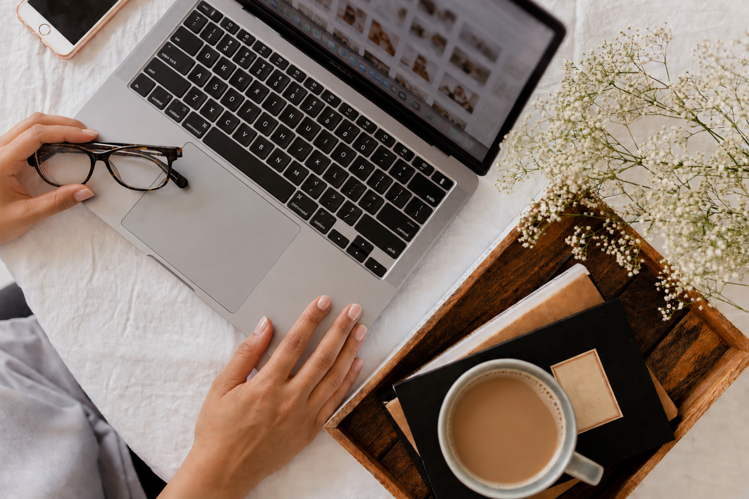 Hands typing on a laptop beside glasses, a coffee cup, a notebook, and fresh flowers on a table.