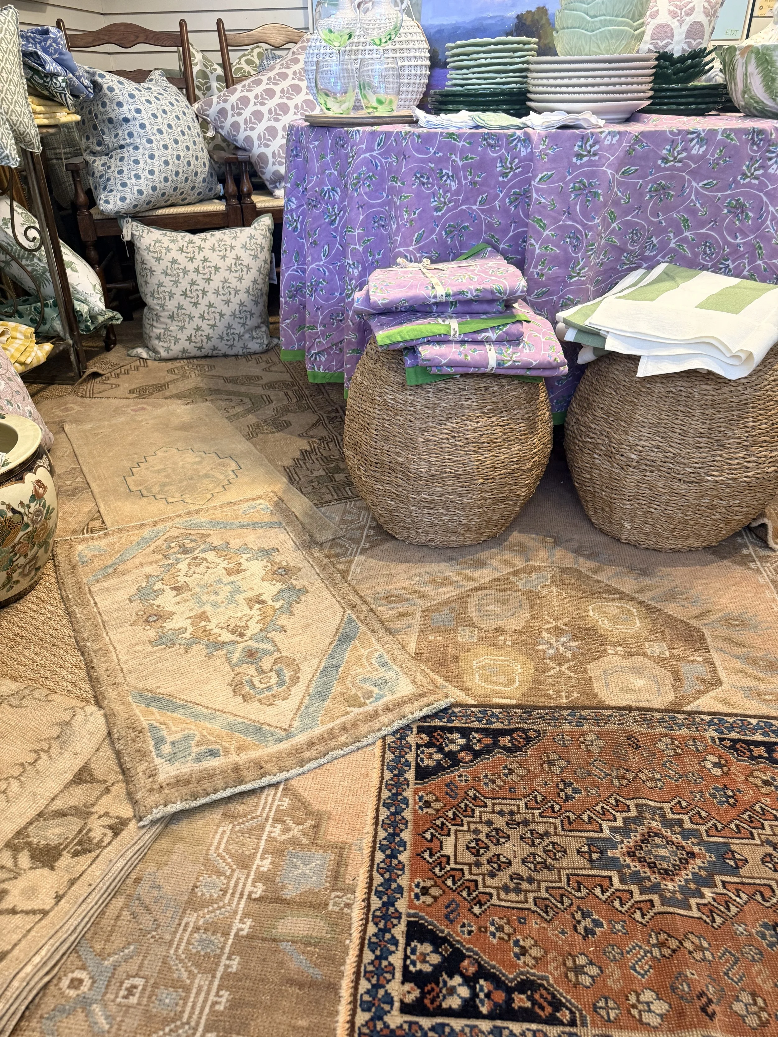 Display of various patterned and solid-colored cushions, plates, bowls, and glassware on a purple covered table with rattan stools in a home decor store.