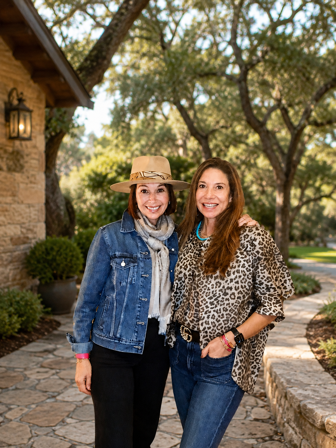 Two women standing outdoors on a stone pathway, smiling, with trees and a house in the background.