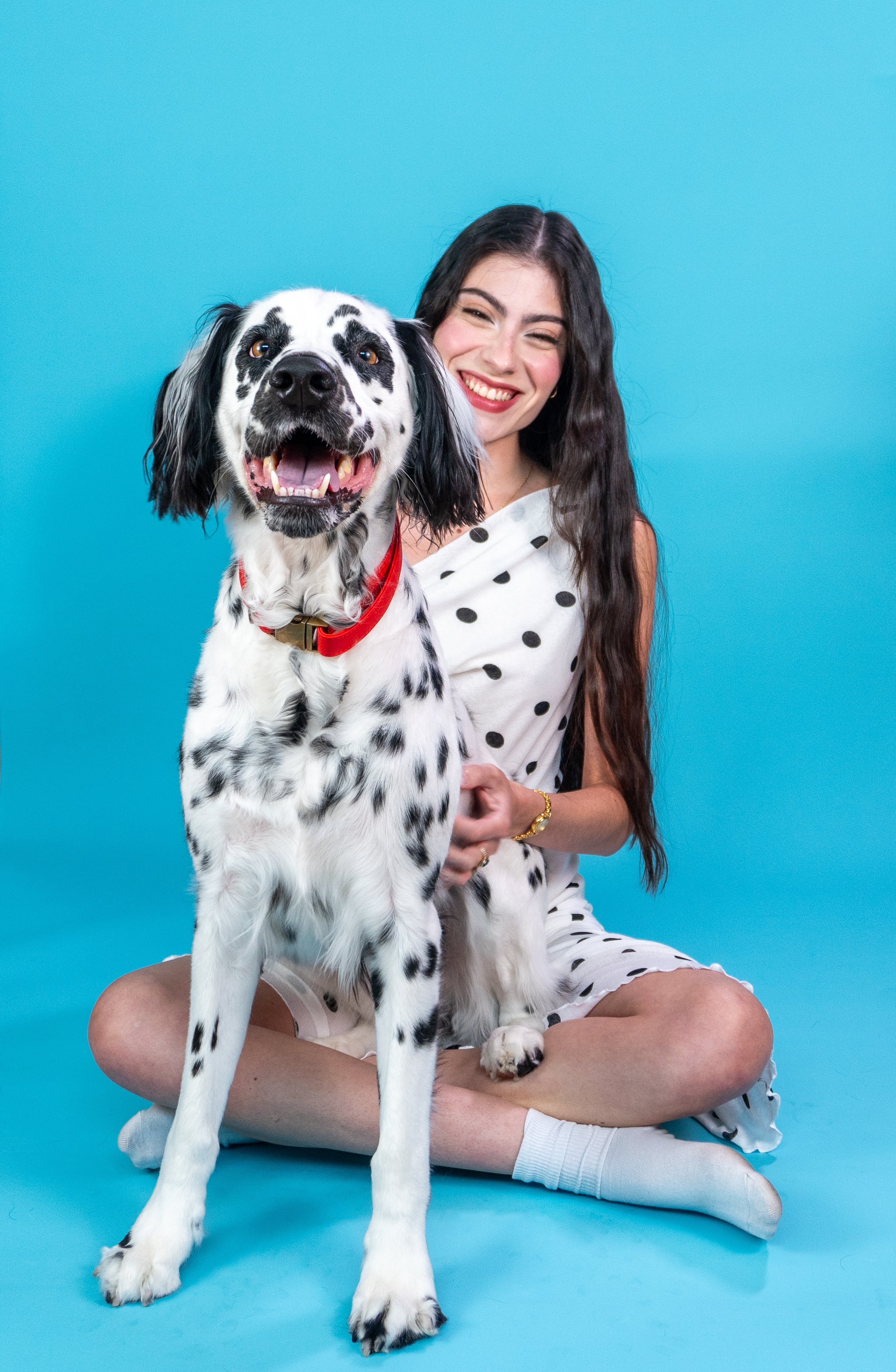 A smiling woman with long, dark hair wearing a white dress with black polka dots, sitting cross-legged on a blue background. There is a large black and white speckled dog with floppy ears, wearing a red collar, standing in front of her.
