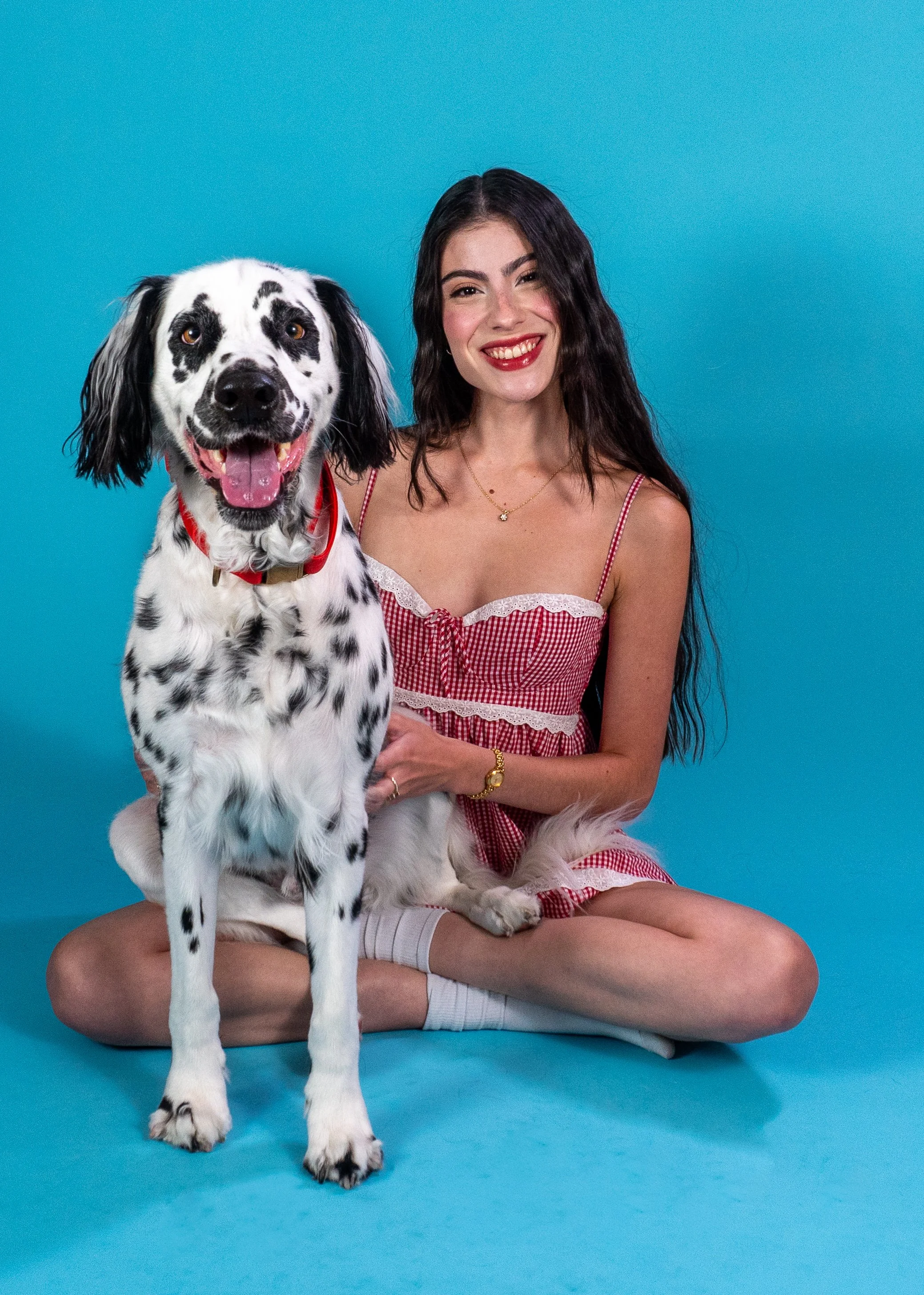 A smiling young woman with long dark hair sitting cross-legged on the floor in front of a bright blue background, holding a large Great Dane dog with a black and white coat and a red collar, both looking at the camera.