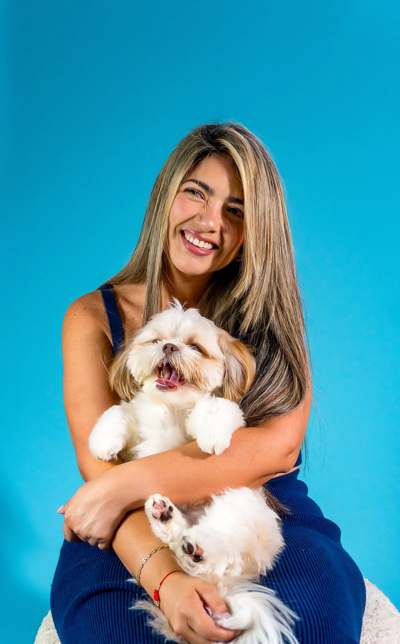 A woman with long blonde hair smiling and holding a sleepy white and tan Shih Tzu dog against a blue background.