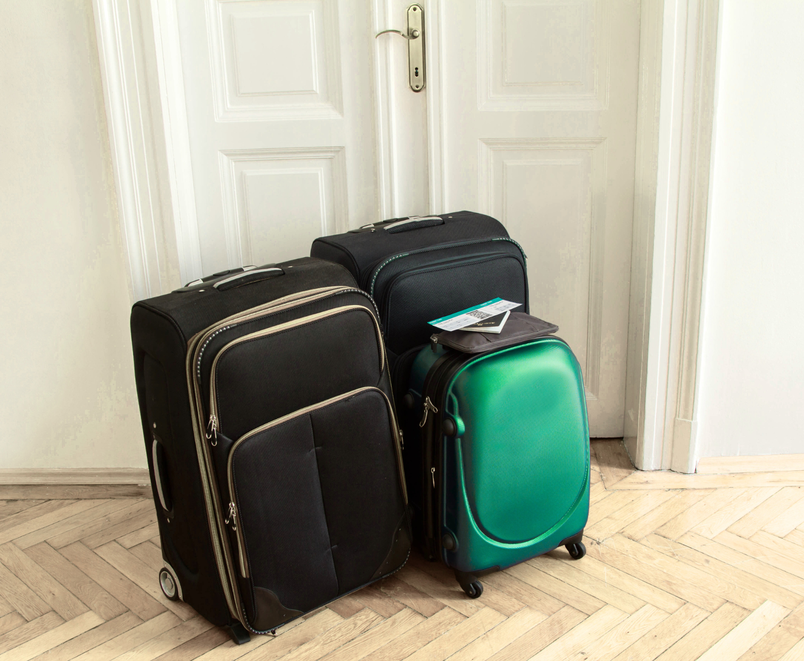 Four suitcases in different sizes and colors - two black, one teal, and one black with a small pouch on top - placed against a white wall and door in a room with wooden parquet flooring.