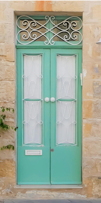 A pair of mint green double doors with white wrought iron scrollwork and a decorative arched iron transom above, set into a sandstone building facade with ivy growing along one side.