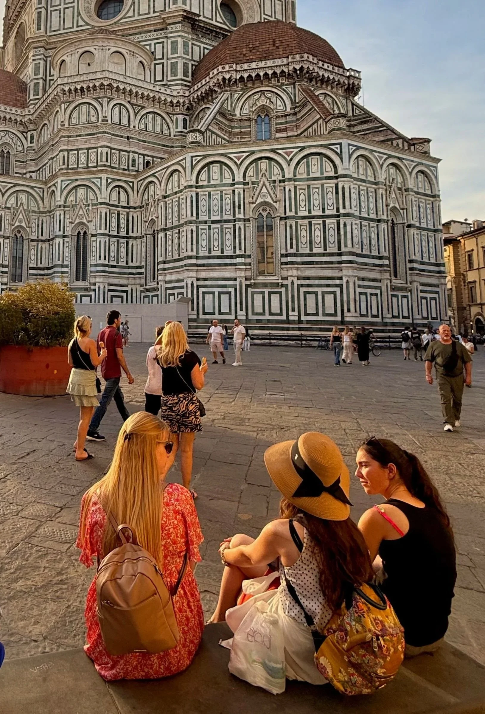 A crowded square in front of the Florence Cathedral, or Cathedral of Santa Maria del Fiore, with people sitting and walking on cobblestone ground during daytime. The cathedral's ornate marble exterior is visible in the background.