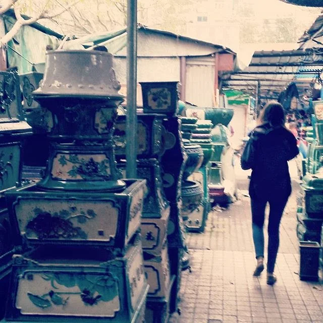 A woman walking through an outdoor market aisle filled with vintage items, including porcelain vases and decorative boxes, on a covered walkway in Zhongshan, China.